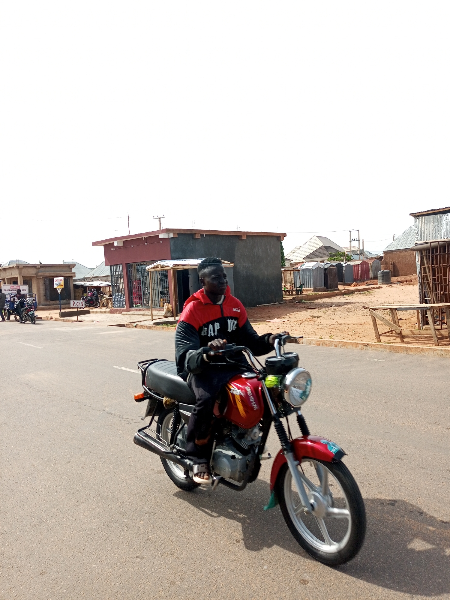 A man rides a red motorcycle down a paved road in Tempure, Nigeria. He is wearing a black and red jacket with "GAP" written on it, black pants, and white sandals. The motorcycle has a silver wheel and a headlight. In the background, there are several buildings, including a red and gray building with shops and a sign that reads "SKYHUB HOTEL." There are also other motorcycles and people visible. The sky is bright and overcast.