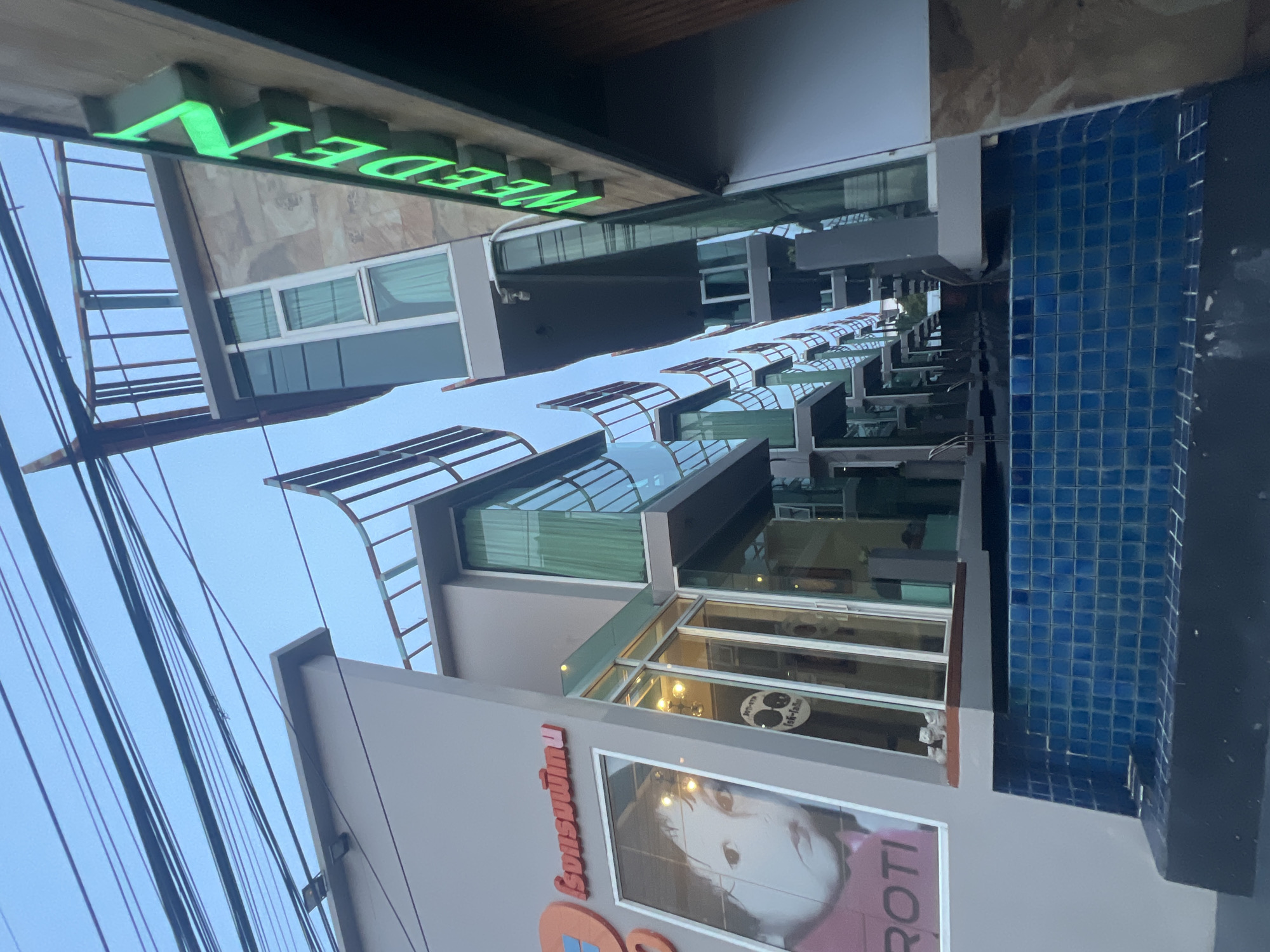 In Ko Samui District, Thailand, the image depicts a low-angle shot of a modern building complex with a swimming pool in the foreground. The architecture features multiple levels with glass facades and balconies, suggesting a hotel or residential property. A neon green sign with the word "WEEDEN" is prominently displayed on one of the buildings. The pool area is tiled in blue. Overhead, a network of electrical wires crisscrosses the sky, indicating an urban or developed area. The overall scene suggests a contemporary commercial or hospitality establishment.