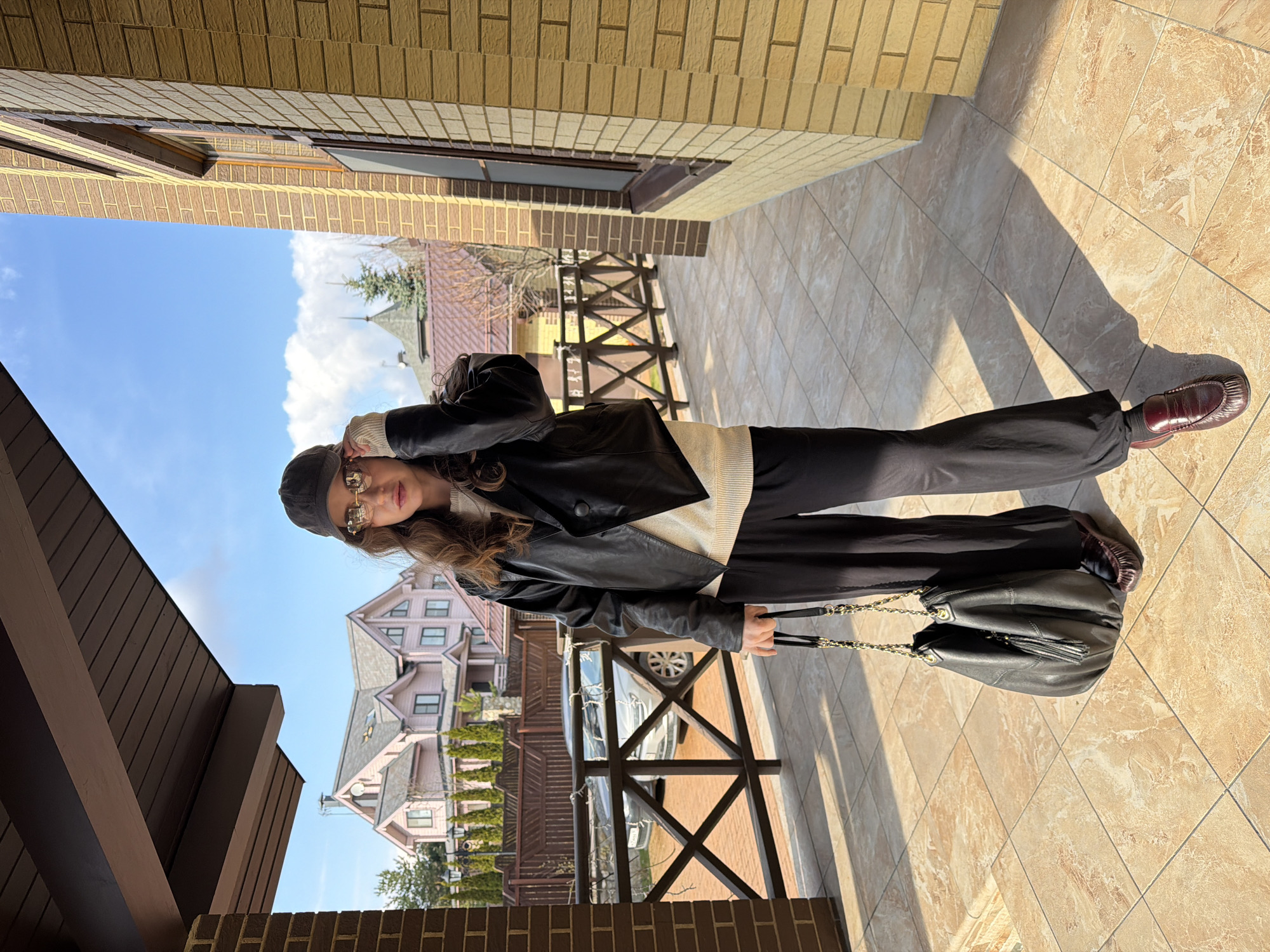 A photograph depicts a person in an outdoor setting, likely a patio or walkway, in what is possibly the city of Mishutkino, Russia. The individual is wearing a black leather jacket, a light-colored sweater, black trousers, and brown loafers. They are also wearing sunglasses and a dark cap, and holding a black leather handbag with gold chain straps.
The background features a building with yellow brick exteriors and a wooden railing, suggesting a residential area. Across the railing, a view of houses with pink exteriors and varying roof designs is visible, indicating a residential neighborhood. The presence of trees and a glimpse of a blue sky with clouds suggests the photo was taken on a clear day. The ground is tiled with light-colored, patterned tiles. The person is partially obscured by the angle of the shot, but their overall posture and attire suggest they are standing and posing for the photograph. A vehicle is parked behind the fence. The overall ambiance indicates a bright, sunny day in a suburban or residential area, possibly in Mishutkino, Russia, given the architectural style of the houses.