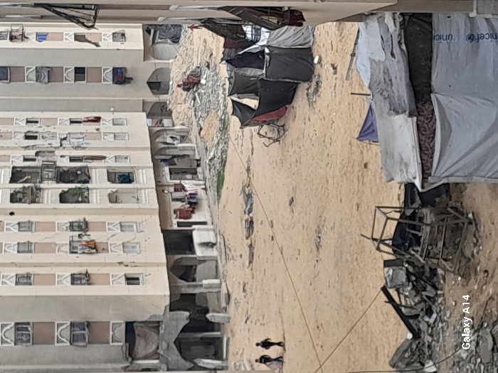 The image captures a devastated urban environment in Khan Yunis, Palestinian Territory. The scene is dominated by a large, open expanse of sandy ground, littered with debris. In the background, several multi-story apartment buildings, light-colored with repetitive window patterns, show extensive damage. Many windows are open or covered with fabric, and clothes hang from balconies and exposed window frames. The lower sections of some buildings appear partially collapsed, with exposed rebar and concrete rubble.
Across the sandy ground in the mid-ground, two dark-clad individuals walk from left to right. Further back, near the base of the buildings, indistinct figures are visible, including one near laundry hanging outside a ground-level opening. To the right of a central pile of rubble, a small figure in red is present.
Scattered across the sandy area are numerous piles of concrete rubble and twisted metal. In the foreground-left, a prominent metal frame structure lies amidst a debris pile. In the mid-ground and foreground-right, several makeshift shelters are constructed from dark and light gray tarpaulins; one light gray tarp bears a "unicef" logo. A bicycle frame or similar twisted metal object is visible near these shelters. Utility lines traverse the scene diagonally across the sandy expanse.