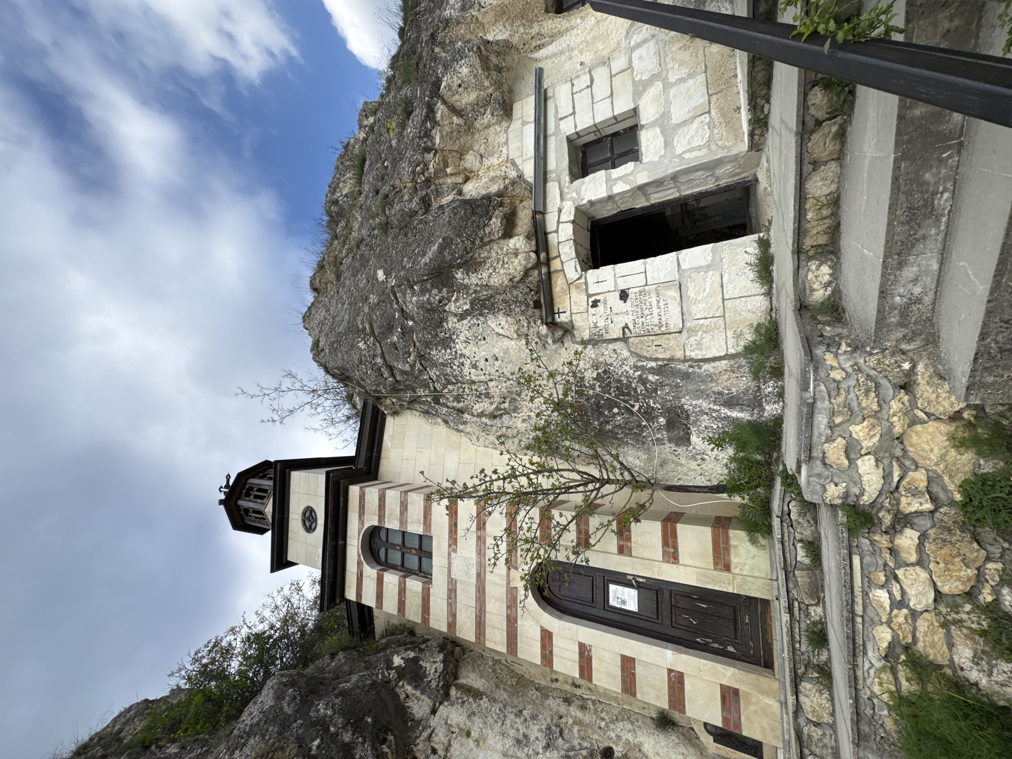 This image depicts the Basarbovo Rock Monastery in Bulgaria, an ancient religious complex integrated into a rugged limestone cliff face under a partly cloudy sky. The scene showcases a fascinating blend of natural rock formations and human-built structures.
In the lower left, a distinct church building stands, characterized by its facade of alternating horizontal stripes of light-colored stone and reddish-brown brickwork. It features a dark, arched wooden entrance door and an arched window with dark framing. A multi-tiered bell tower, topped with a cross, rises from the roof, silhouetted against the bright sky. Sparse trees with budding branches grow around the church, adding a touch of natural vitality.
To the right and higher up the cliff, several structures are carved directly into the rock or built as facades for cave openings. These are constructed from light-colored, neatly cut stone blocks, featuring rectangular windows and larger darkened openings. A black metal handrail runs alongside a concrete path or staircase, ascending the cliff and indicating access to the various levels of the monastery.
A notable detail is a Cyrillic inscription on a stone plaque embedded in the wall of one of the upper monastic cells. Partially legible text includes "ИИС ХС" (Jesus Christ), a cross symbol, "МОНАХА ХРИСАНТ" (Monk Hrisant), and dates "1910/1938 Г." This inscription likely commemorates a monk or an event within those years.
The image captures the monastery during daytime, with ample natural light. The weather is partly cloudy, featuring patches of blue sky interspersed with white clouds. No people are visible, lending a sense of timelessness and peacefulness to this historical and spiritual site. The overall setting highlights the ingenuity of early monastic communities in utilizing natural caves for their dwellings and places of worship.