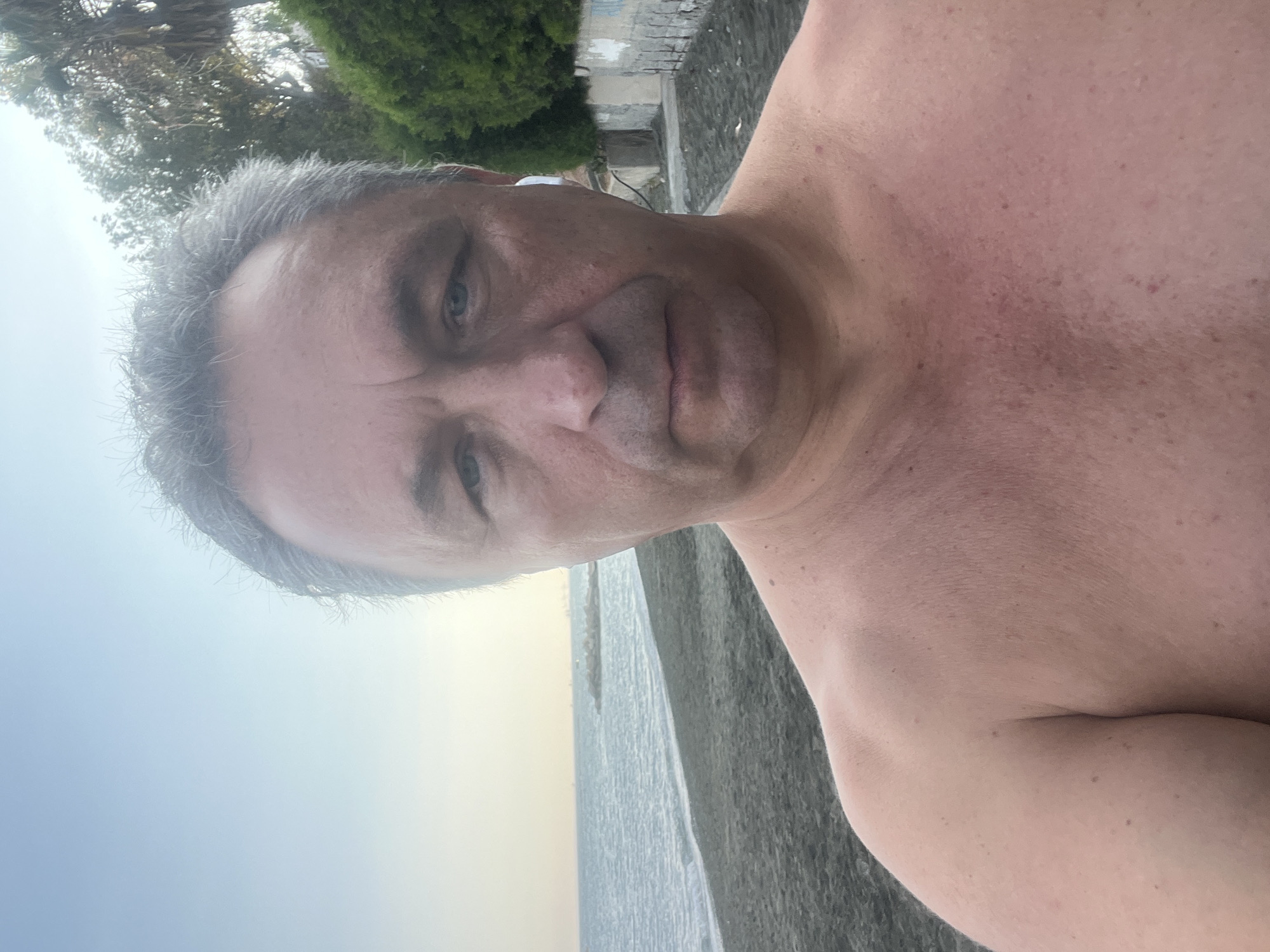 A shirtless man with graying hair and blue eyes takes a selfie on a beach in Agios Tychona Community, Cyprus. The man is wearing white earbuds in his right ear. The background shows a dark sand beach, the sea, and some trees and a wall. The sky is a pale blue.