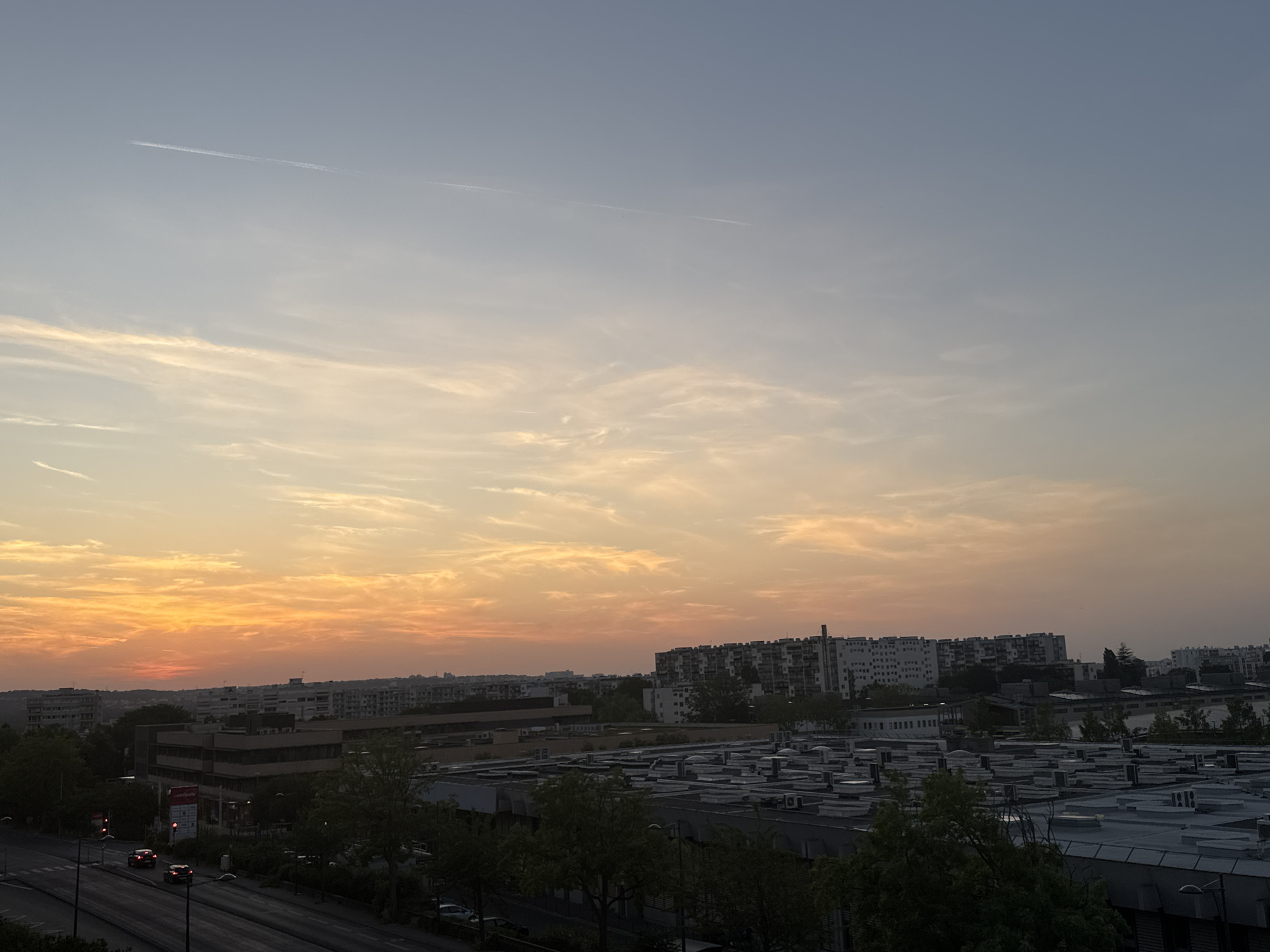 This is a wide shot of a city skyline at sunset. The sky is a mix of soft blues, grays, and oranges, with wispy clouds stretching across the horizon. The sun is low in the sky, casting a warm glow over the scene. In the foreground, there are several buildings, including some low-rise industrial-looking structures with flat roofs and some taller apartment buildings further in the distance. Trees line the streets and are scattered amongst the buildings. A road with a few cars is visible on the left side of the image. A single contrail from an airplane is visible in the upper left corner of the sky. The overall mood of the image is peaceful and serene, capturing a quiet moment as day transitions into night. There is no visible text in the image.