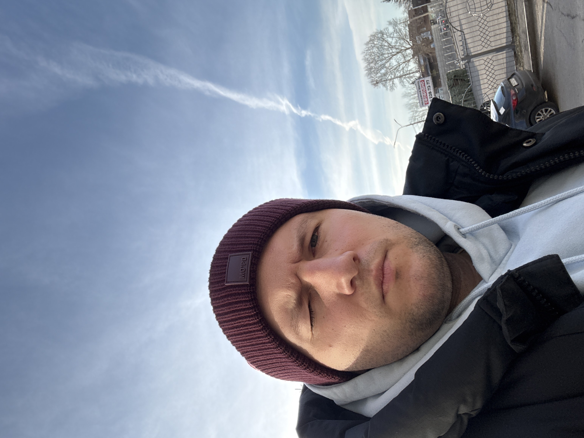 A close-up, selfie-style photograph depicts a man wearing a maroon knit cap, a light gray hoodie, and a black puffer jacket. He appears to be outdoors on a sunny day, with a mostly clear blue sky featuring a prominent contrail. In the background, blurry details suggest a cityscape, including a tree, a fence, a parked car, and a sign with Russian text. According to the text on the sign, the location is in the city of Krasnoyarsk, Russia, near 226-72-77.