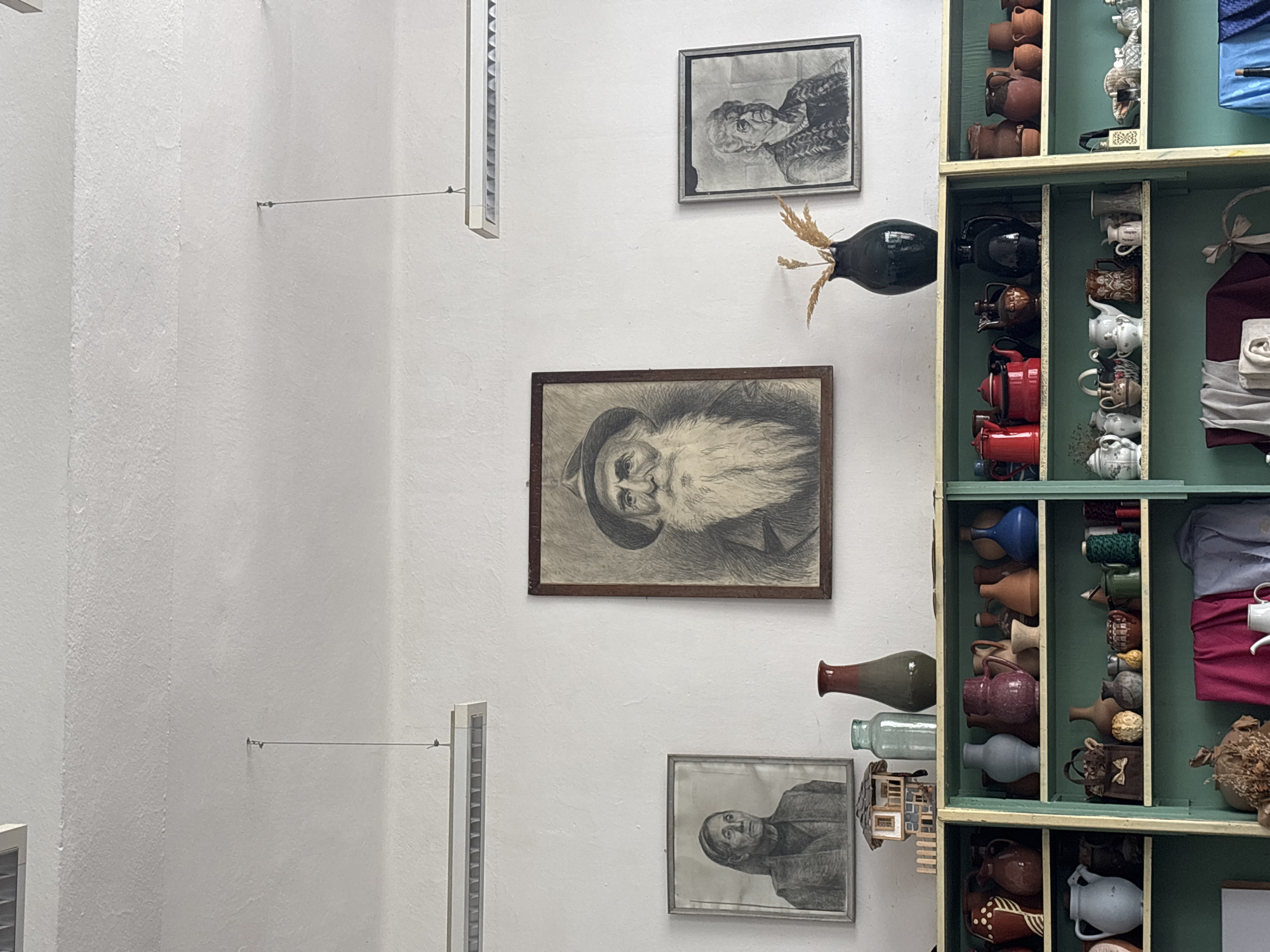 The scene depicts an interior wall, likely in a building in Troyan, Bulgaria. The wall is white and textured. It features three framed portraits in charcoal or pencil. From top to bottom: an older man with glasses, a bearded man with a hat, and another older man. All the men appear to be facing left. To the right of the portraits is a wooden display case, painted green and filled with a variety of pottery, vases, teapots, cups, and fabric items. A vase with dried wheat is placed next to the display case. Two window shutters hang on the wall, and thin wires run along the wall horizontally, connecting the shutters to other objects.