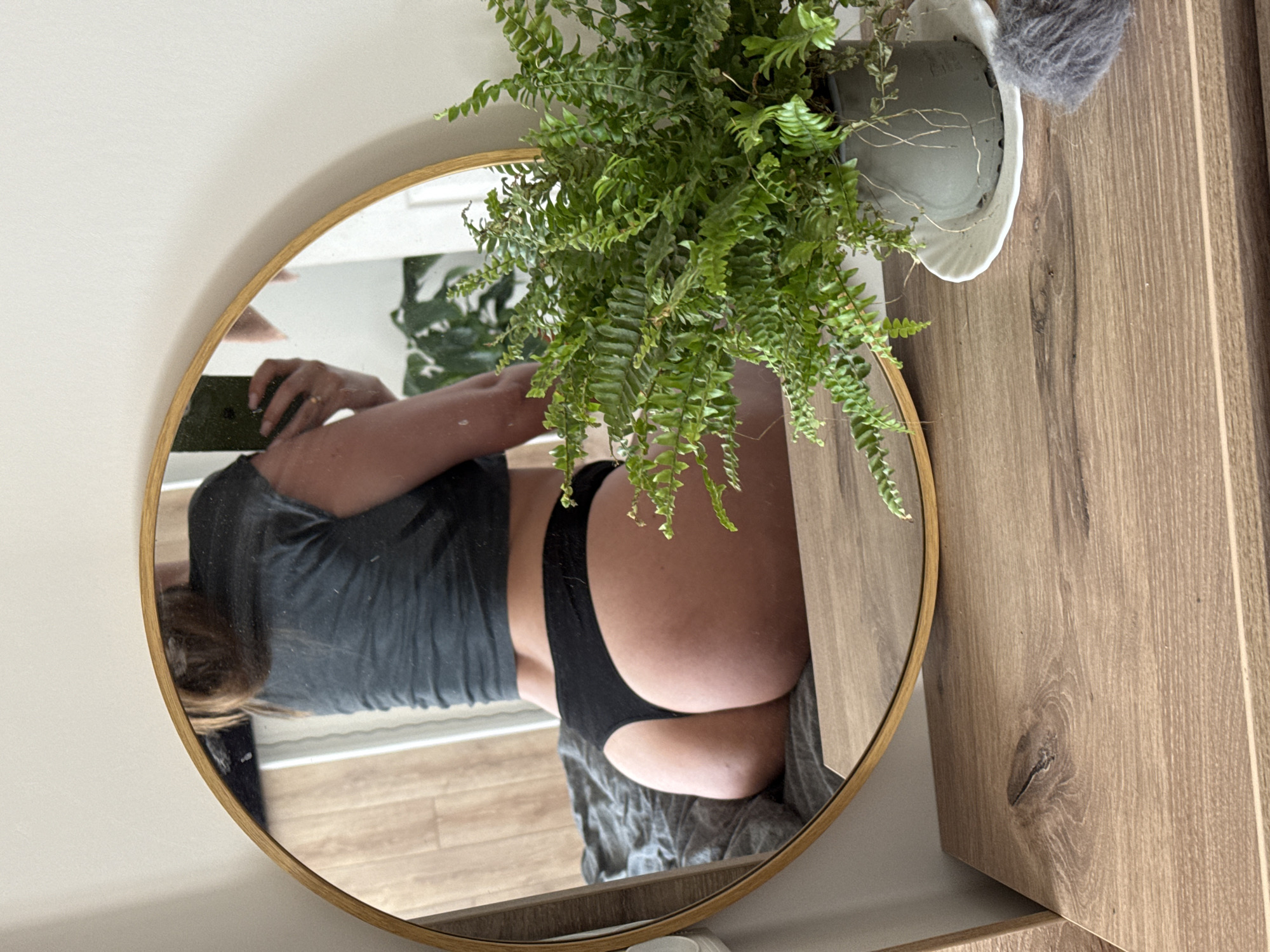 A woman is taking a mirror selfie in a bedroom. The woman is wearing black underwear and a grey t-shirt. She is sitting on a bed with grey sheets. There is a round mirror with a gold frame in front of her. A fern plant in a grey pot is on a wooden dresser next to the mirror.