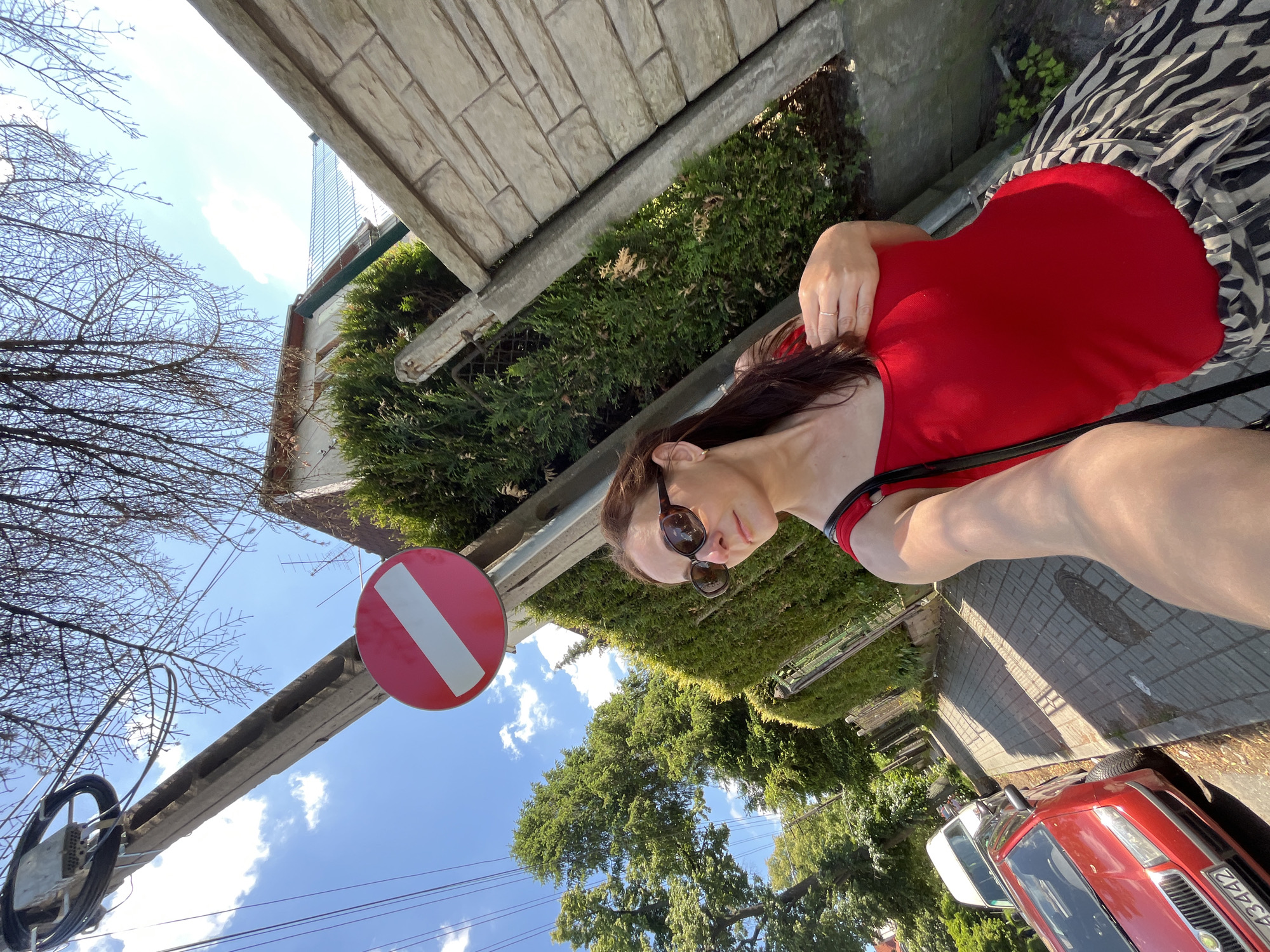 A woman is taking a selfie on a sidewalk in a residential area. She is wearing a red top and a patterned skirt, sunglasses, and a ring on her finger. Behind her, there is a hedge, a stone wall, a traffic sign, a utility pole with wires, and a street with cars parked on it. The sun is shining, and there are clouds in the sky.