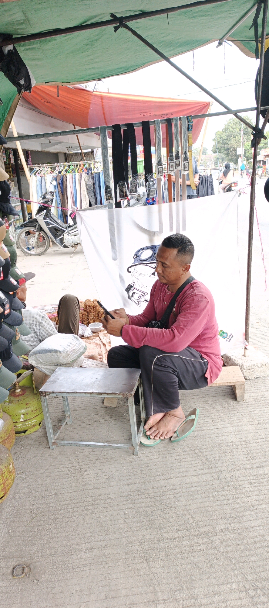 A middle-aged Indonesian man sits on a wooden stool in a roadside market stall in Kasiyan, Indonesia, engrossed in his mobile phone. He is wearing a red long-sleeved shirt and black pants, with light green flip-flops. A small, metallic grey stool is positioned in front of him. Behind him, a white banner with a black graphic design of a helmeted head is displayed. The stall is covered by a green and orange tarpaulin. Various merchandise, including belts hanging on display, clothing on racks, and gas cylinders, are visible in the background. A motorcycle is parked nearby. The ground is paved with concrete. The scene depicts a typical street market environment.