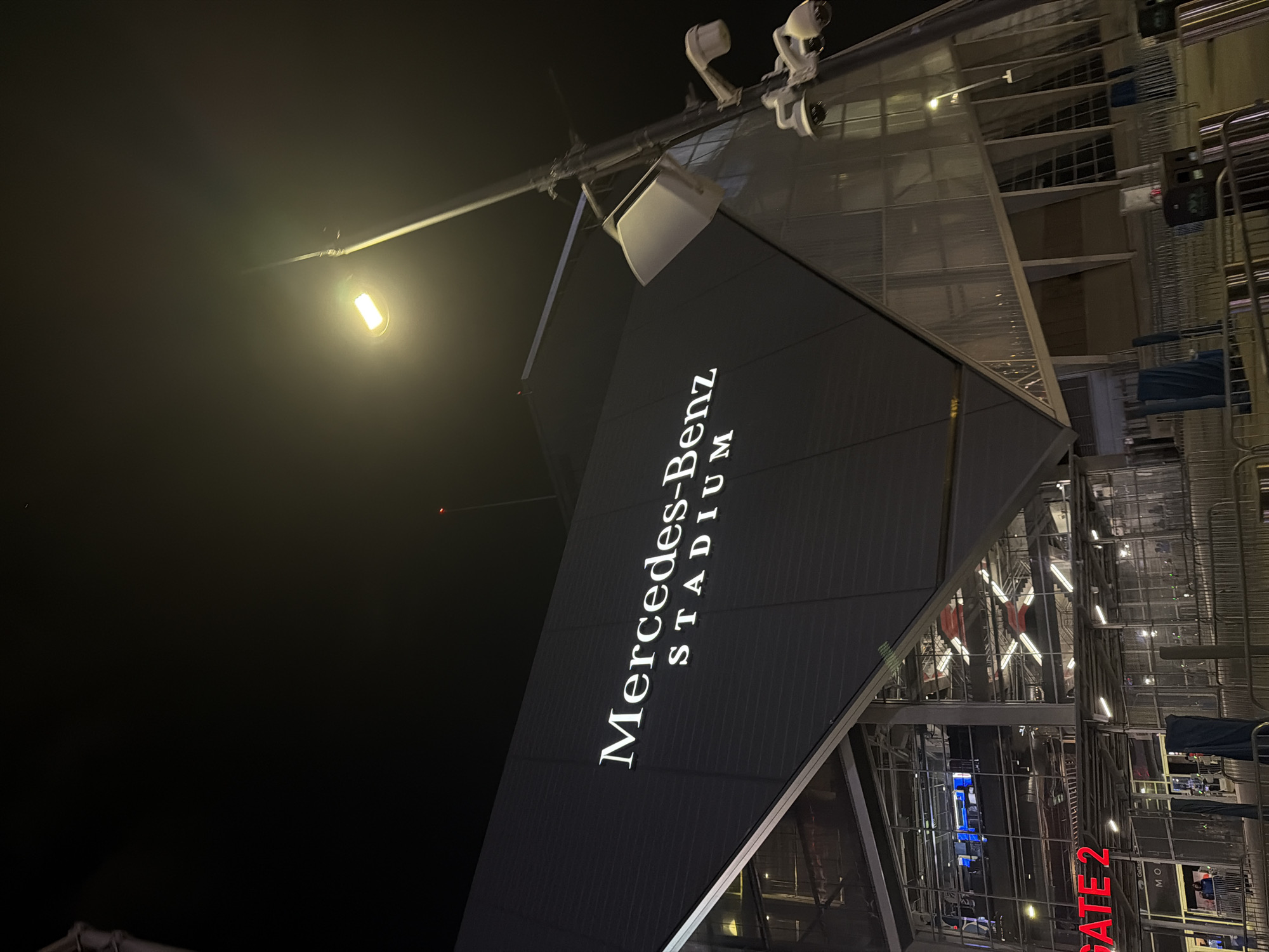 The image captures an exterior view of Mercedes-Benz Stadium in Atlanta, United States of America, at night. The stadium's distinctive angular architecture is prominent, featuring the illuminated "Mercedes-Benz STADIUM" signage in a vertical arrangement. A streetlight casts a bright glow above, and surveillance cameras are mounted on a structure near the stadium's upper section. The right side of the frame reveals elements of the stadium's exterior, including metallic structures, lighting, and signage for "GATE 2" in red. The overall scene suggests an urban environment, with the focus primarily on the stadium's architectural features and lighting.