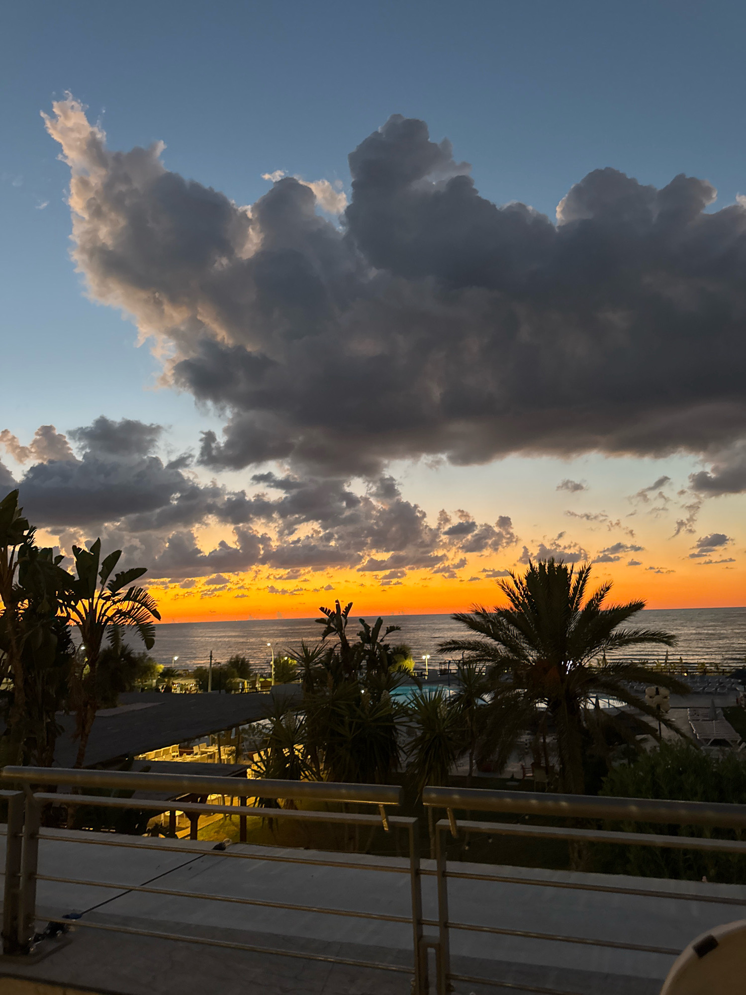 The image shows a sunset over the sea in Isola delle Femmine, Italy. The sky is filled with dramatic clouds in shades of gray, white, and orange. The sun is low on the horizon, casting a warm glow over the water. Palm trees and other vegetation are silhouetted against the colorful sky. In the foreground, a balcony railing overlooks the scene. There are no people visible in the image. The scene appears to be a resort or hotel with outdoor seating and a swimming pool.