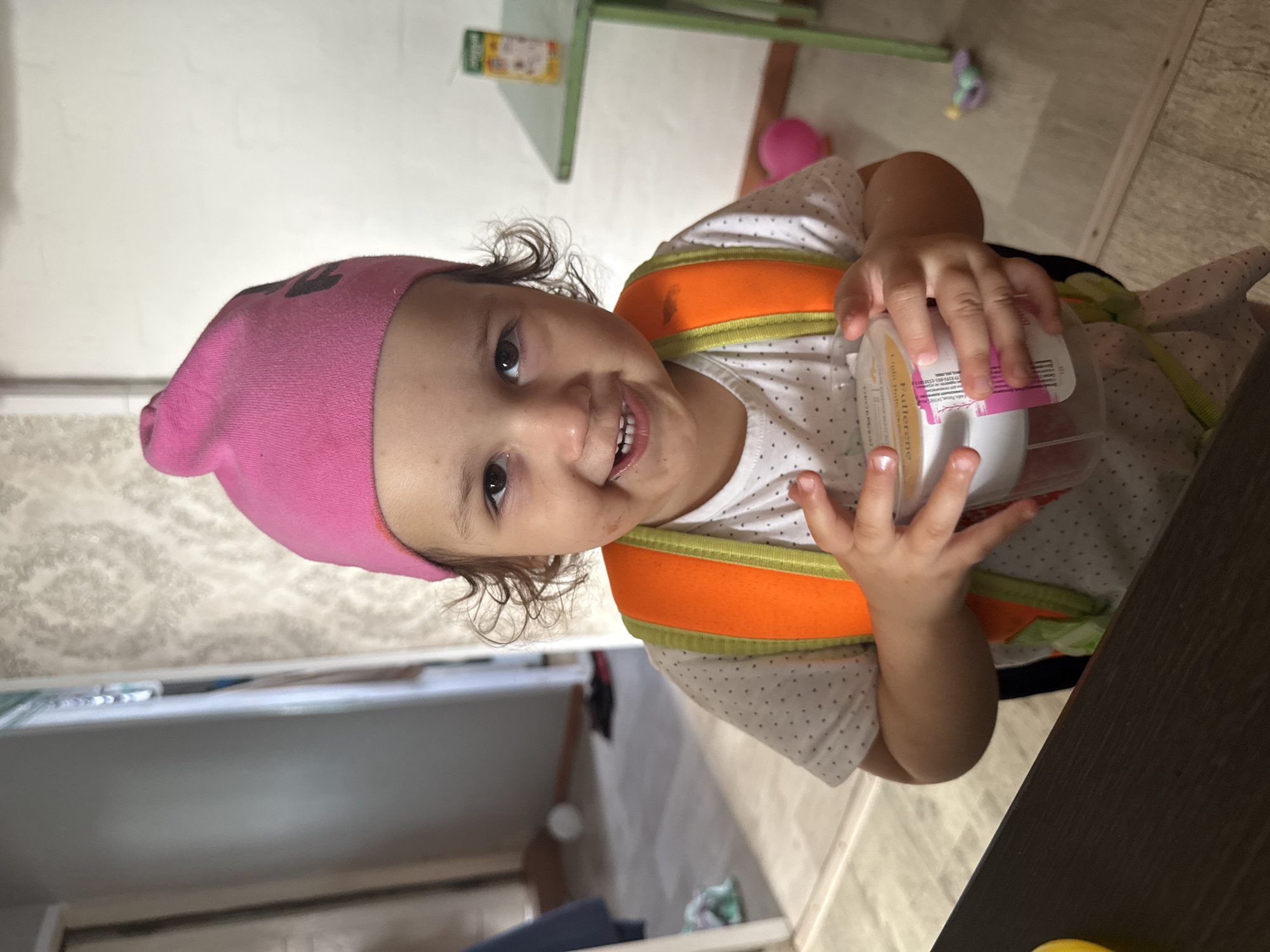 This portrait-oriented photo, taken in Уральское, Russia, captures a cheerful young child indoors. The child, likely a toddler or preschooler, has dark curly hair and a wide, happy smile, looking directly at the camera. They are wearing a bright pink beanie, a white short-sleeved top with small dark polka dots, and a distinctive orange and lime green harness or vest over the top.
The child is holding a clear plastic container with a white interior, possibly a snack or dessert cup. On the container's label, partial words like "Pâtisserie" and numbers including "13:09" are visible, along with other small, indistinct text and a barcode.
The indoor setting features a light-colored wall in the background, with a light green shelf holding a small item. A dark wooden table or counter surface is visible to the right and bottom. The floor appears to be light-colored laminate or tile. A pink ball and a small toy are also visible in the background. The child's happy expression suggests a joyful moment, likely within a home environment.