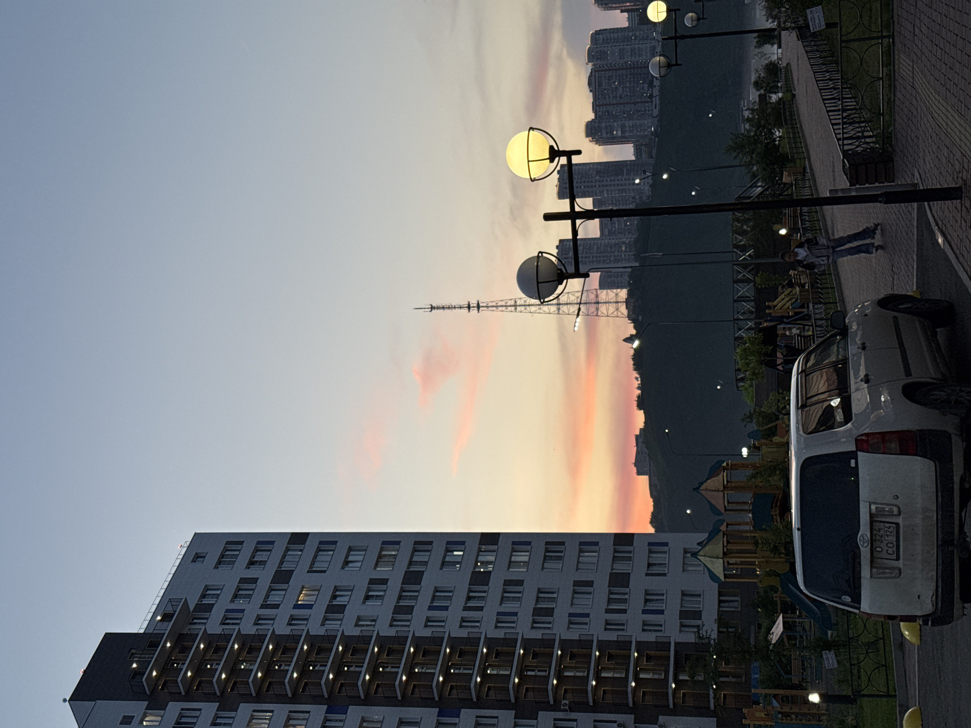 The image depicts a dusk scene in the city of Krasnoyarsk, Russia. In the foreground, a white minivan is parked on asphalt. To the left, a modern, tall apartment building with multiple balconies and illuminated windows dominates the frame. A children's playground with colorful structures is visible near the base of the building. A paved walkway with lampposts runs horizontally across the middle ground, where a young person is rollerblading. In the background, a cityscape with various buildings and a prominent radio tower is silhouetted against a sky filled with soft pink and orange clouds, indicative of sunset. The environment appears to be a residential area with recreational facilities, likely overlooking a body of water or a valley, given the hilly terrain and distant buildings.