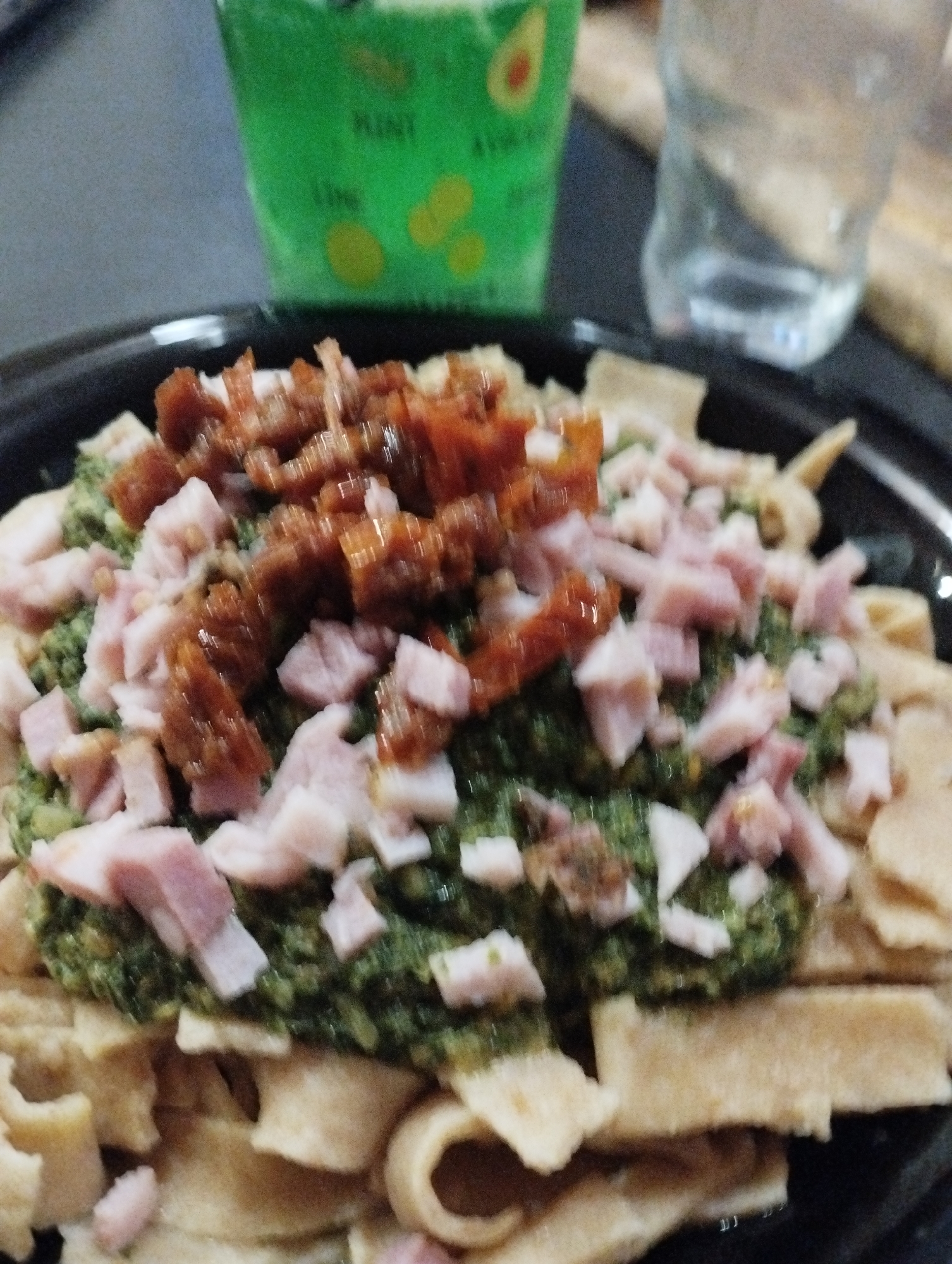 A close-up photograph shows a black bowl filled with pasta topped with green pesto, diced ham, and chopped sun-dried tomatoes. Behind the bowl, slightly out of focus, is a green container with avocado designs and a glass of water. The background suggests an indoor setting, possibly a restaurant or cafe. Given the instructions, the location would be in Burgas, Bulgaria. The image focuses primarily on the food presentation, with other elements in the background for context.