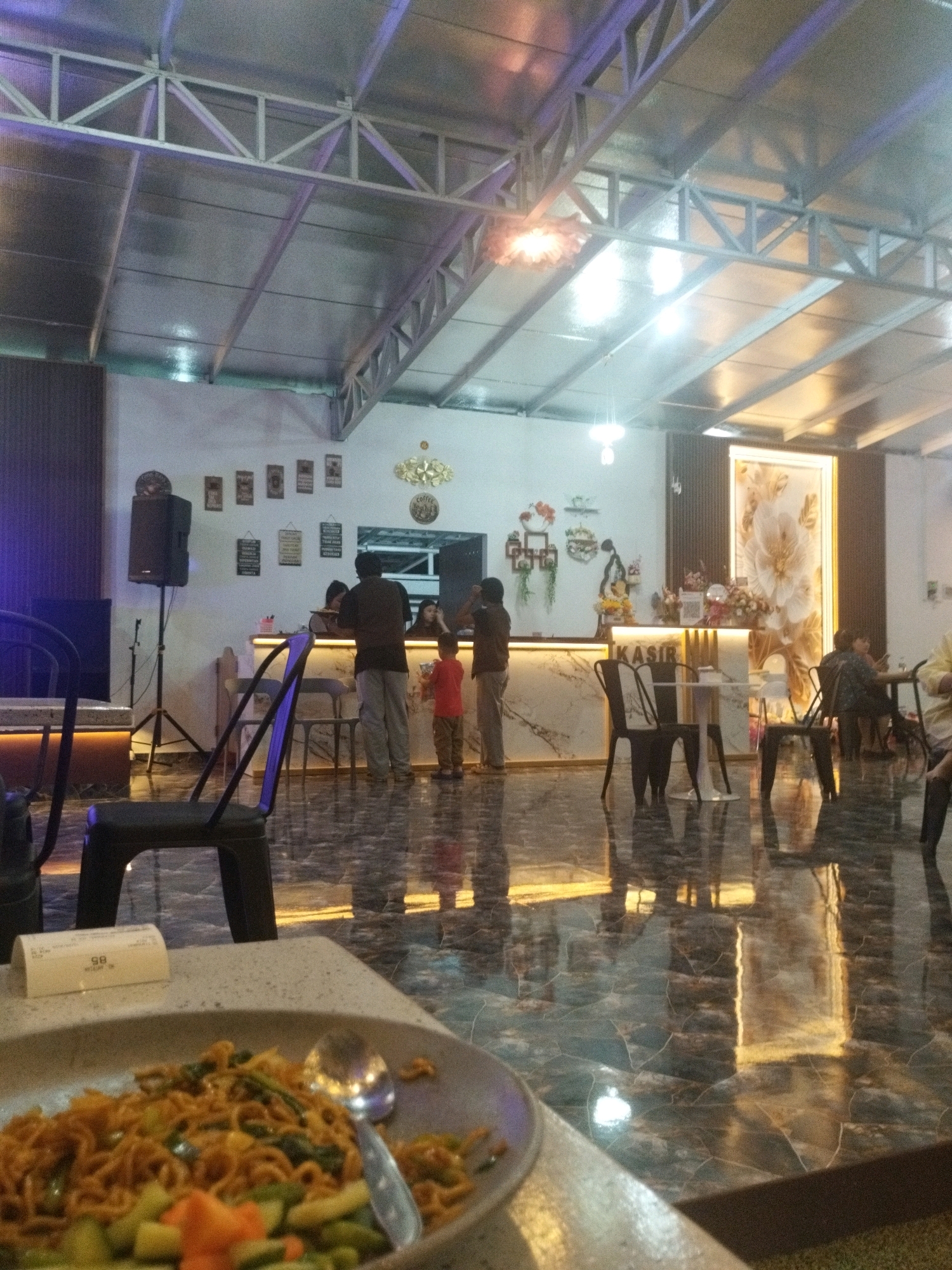 This image depicts the interior of a restaurant or cafe, likely located in Sungailiat, Indonesia, during the evening. The scene is characterized by its modern, somewhat industrial aesthetic, with a prominent metal truss ceiling and reflective, dark marble-patterned flooring.
In the foreground, a plate of food, appearing to be noodles with vegetables, rests on a table. A spoon is visible in the plate. To the left, a black chair is positioned in front of another table.
The midground features a counter area with "KASIR" (Indonesian for "cashier") illuminated on it. Several people are present around the counter. A man in a dark shirt and grey pants stands with his back to the camera, interacting with someone behind the counter. A young boy in a red shirt and khaki pants stands near him, facing the counter. Another person, possibly a woman, is seated behind the counter. Further to the right, two chairs are placed at a small table with two individuals seated.
The walls are decorated with framed pictures and a large floral artwork. A speaker system is visible to the left, mounted on a stand. Lighting fixtures are integrated into the ceiling and behind the counter, creating a warm ambiance. The overall impression is of a busy, well-lit establishment.