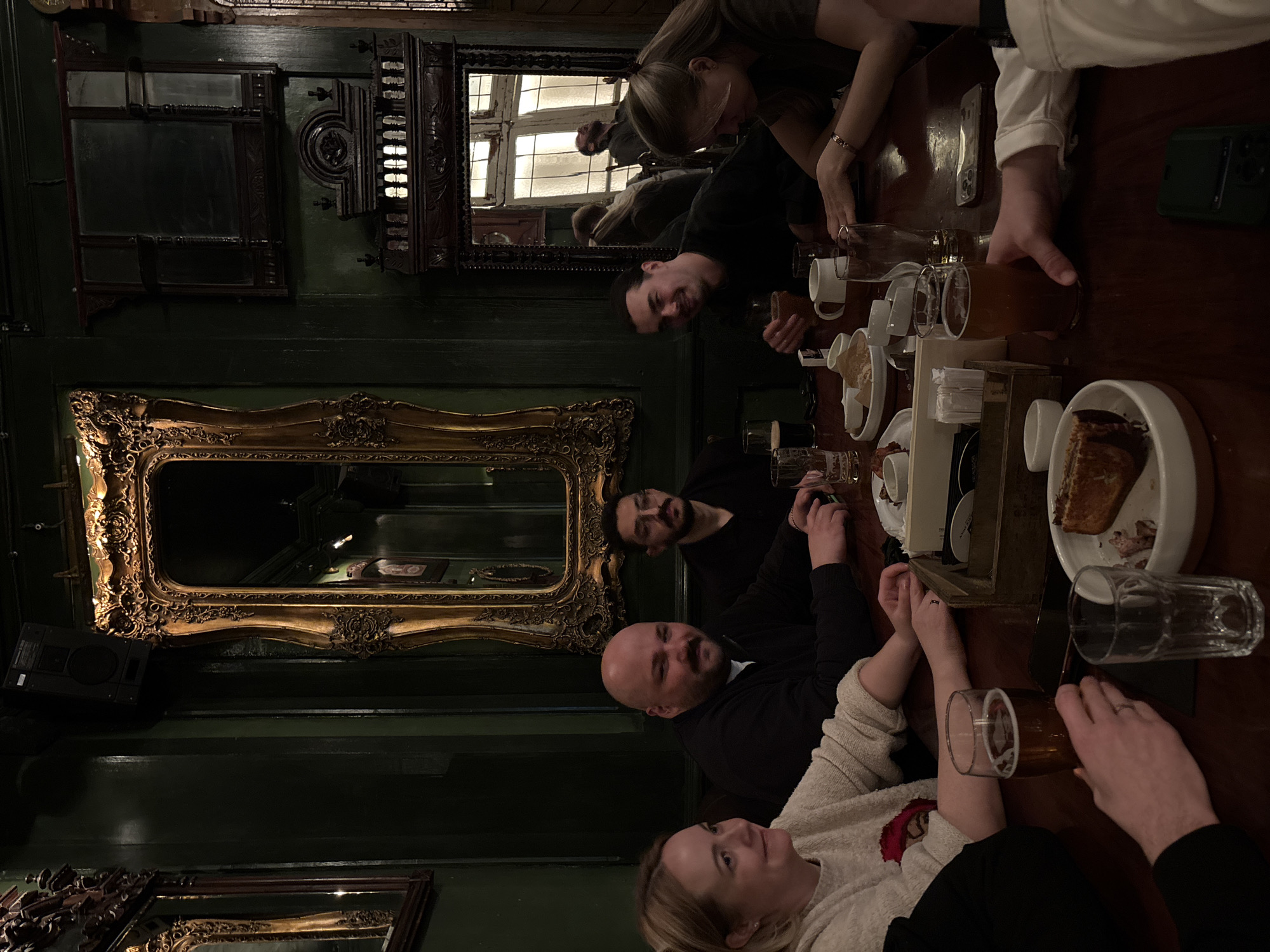 The image depicts a group of six people seated around a wooden table in what appears to be a pub or restaurant. The setting has dark green walls adorned with ornate mirrors. The table is covered with drinks, mugs, plates, and food. People are interacting and socializing. The location is Unknown, Unknown.