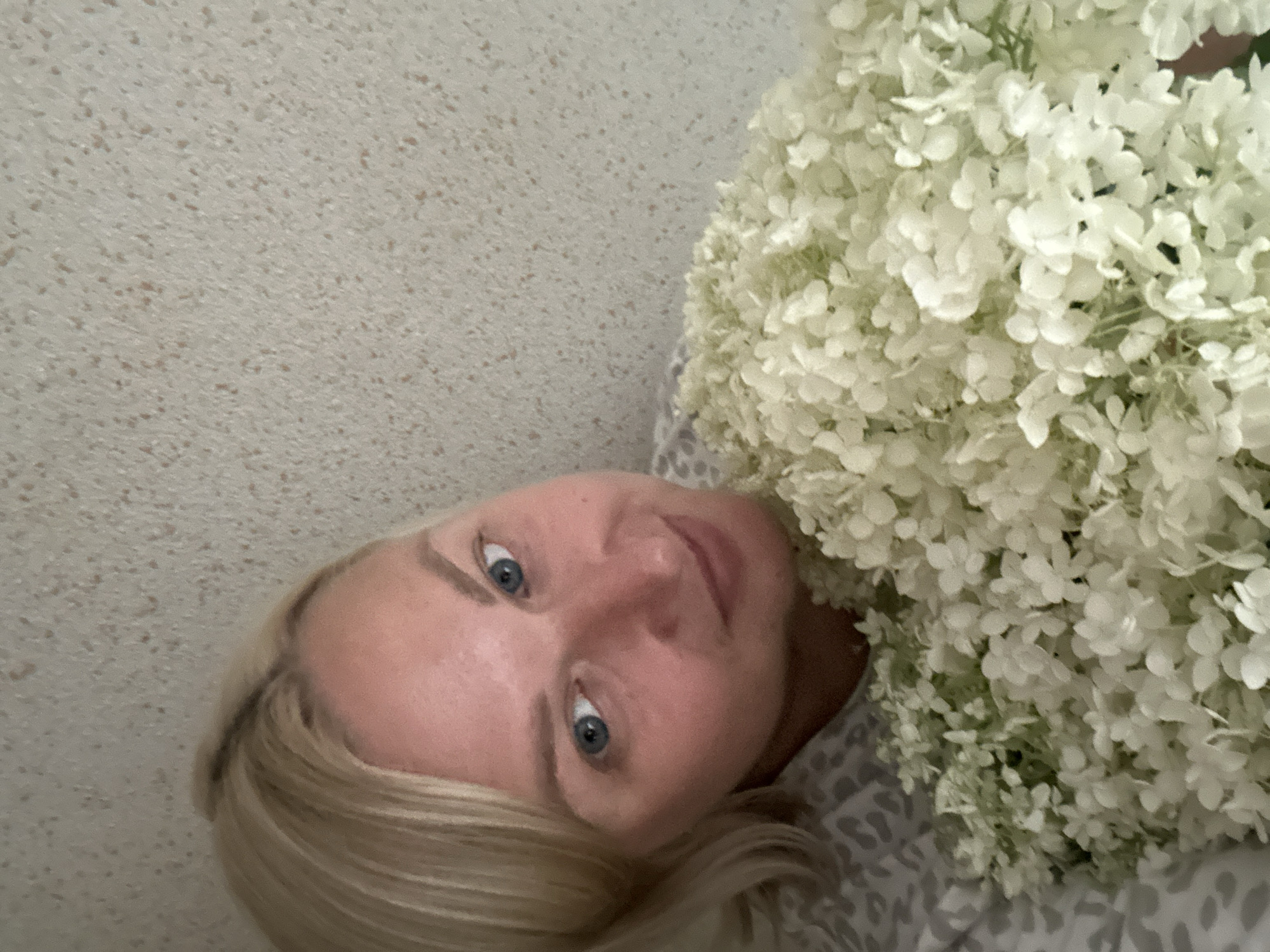 A woman with short blonde hair and blue eyes is lying down, holding a large bouquet of white flowers. She is wearing a patterned top. The background appears to be a textured wall. The image is a selfie, taken from a close-up perspective. The location in Chaikovsky, Russia, is not discernible from the image.