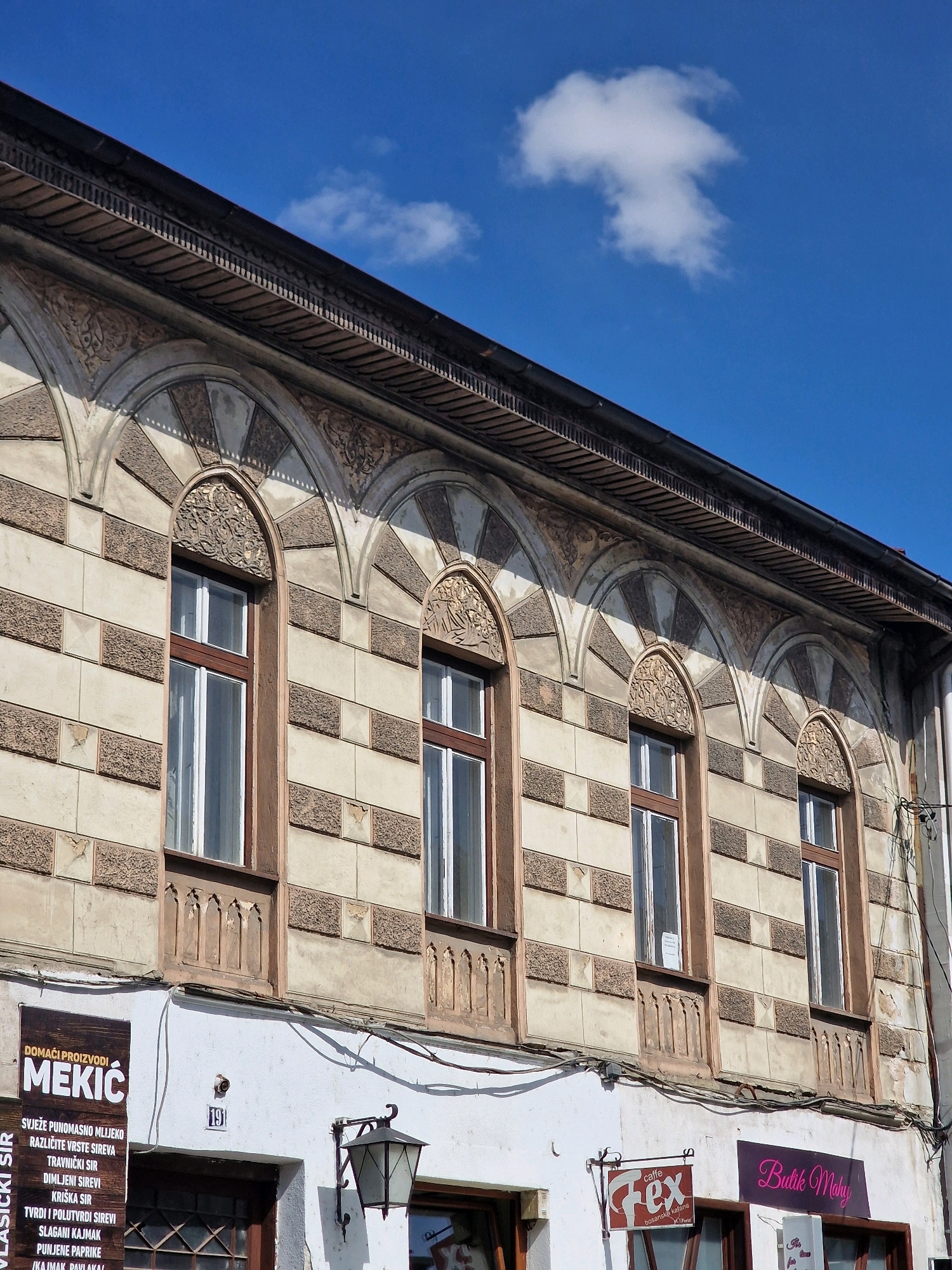 This is a photograph of the facade of a building in Travnik, Bosnia and Herzegovina, under a clear blue sky with a single white cloud. The building has a distinctive architectural style featuring a series of arched windows with decorative stonework above them. The facade is primarily light-colored with a pattern of darker rectangular blocks. Below the windows, there are storefronts with signs. On the left, a vertical sign with white text on a brown background reads "DOMAĆI PROIZVODI MEKIĆ" and lists various dairy products. Next to it, a sign for "caffe Hex" hangs. Further to the right, a magenta sign reads "Butik Maky". Electrical wires are visible running along the building's exterior. An ornate streetlamp is mounted to the wall.