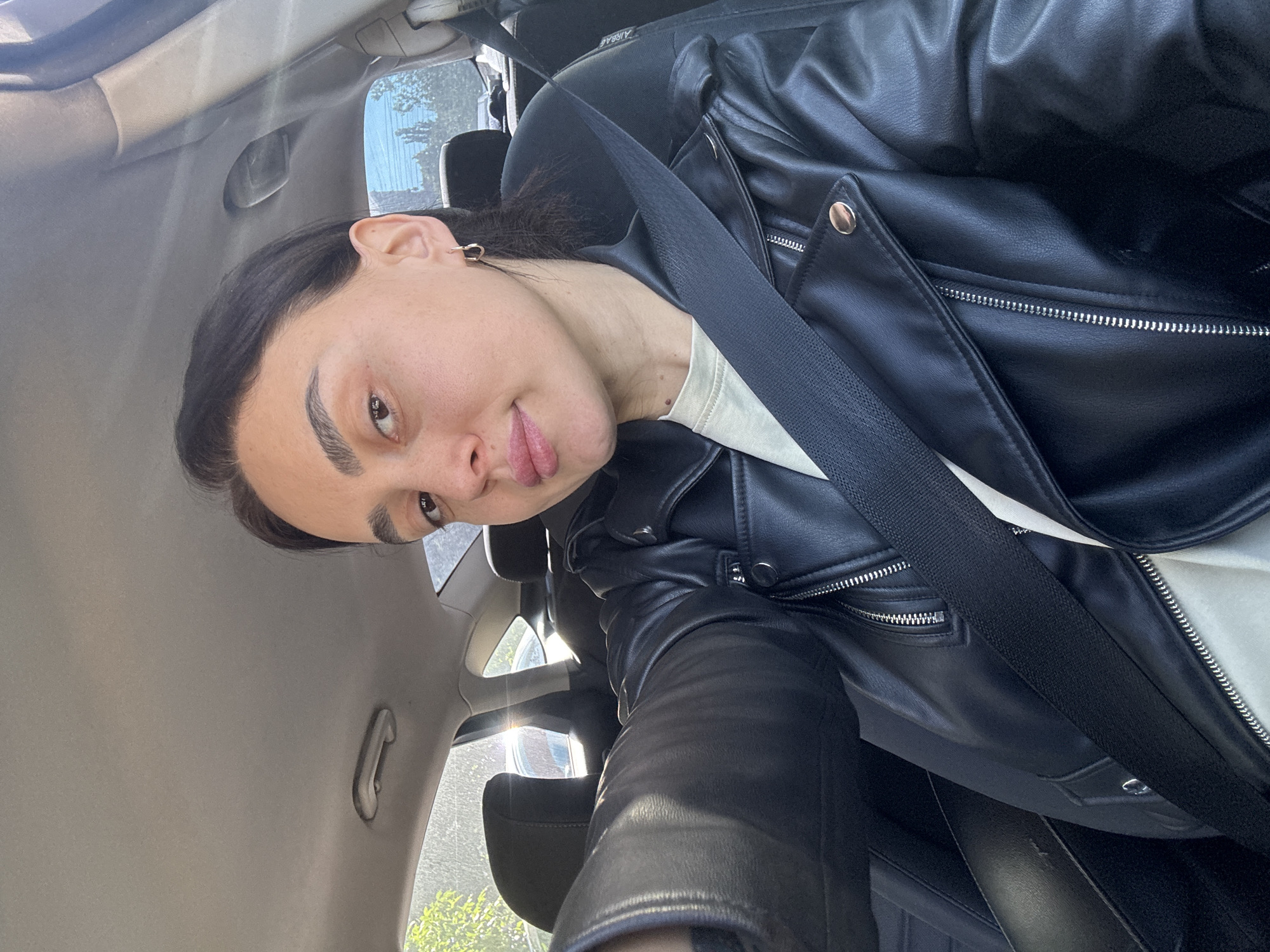 A person with fair skin, dark eyebrows, and short, dark hair is wearing a black leather jacket and a seatbelt in a car. The car's interior is visible, including the ceiling, door, and seat. Outside the car, there are hints of trees and buildings, suggesting a city environment. The image suggests the person is inside a vehicle, possibly a taxi or private car, traveling in Tbilisi, Georgia.