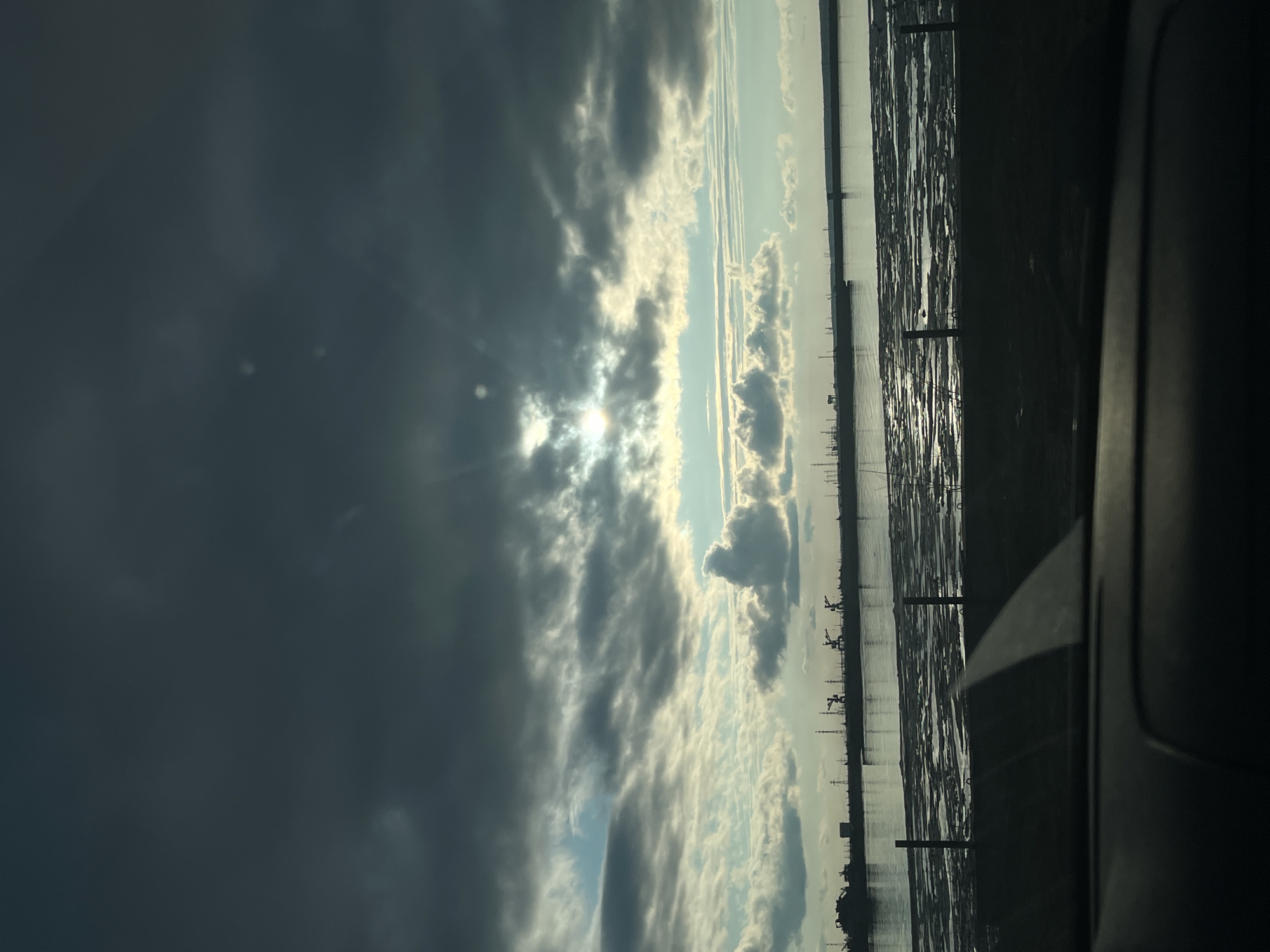 The image presents a dramatic sky over a wide body of water, likely the Kama River in Chaikovsky, Russia, viewed from a vehicle. The sun, partially hidden by dense, dark gray clouds on the left side of the frame, emits bright rays that break through the cloud cover, causing a noticeable lens flare and illuminating sections of the sky with a brilliant white glow. The clouds vary in density and color, ranging from heavy dark formations to lighter, more ethereal wisps, revealing patches of lighter blue sky.
The right side of the image is dominated by the water, which reflects the bright sky and sun, creating a shimmering, almost textured surface. Along the distant horizon line on the right, numerous dark silhouettes of what appear to be industrial cranes or other structures are visible, suggesting a port or industrial waterfront. The extreme right edge of the frame shows a dark, blurry shape consistent with a car's window frame or interior, indicating the photograph was taken from within a vehicle.
The scene captures a dynamic weather condition, likely during late afternoon or early morning, characterized by striking sunbeams through a cloudy sky. No discernible text is visible in the image.