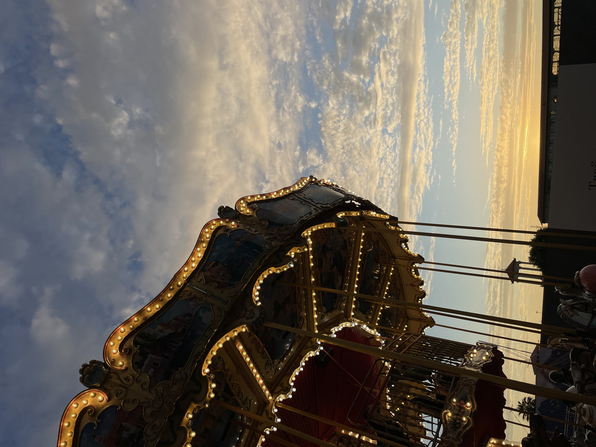 A carousel is illuminated with lights at dusk, set against a cloudy sky in Sochi, Russia. The carousel, painted in dark blues and golds, is ornately decorated and features various animal figures, some of which are partially visible. The sky displays streaks of orange and yellow from the setting sun, with scattered white clouds. In the background, a few buildings and a distant structure with a dome are silhouetted against the fading light. The overall scene evokes a sense of twilight amusement.