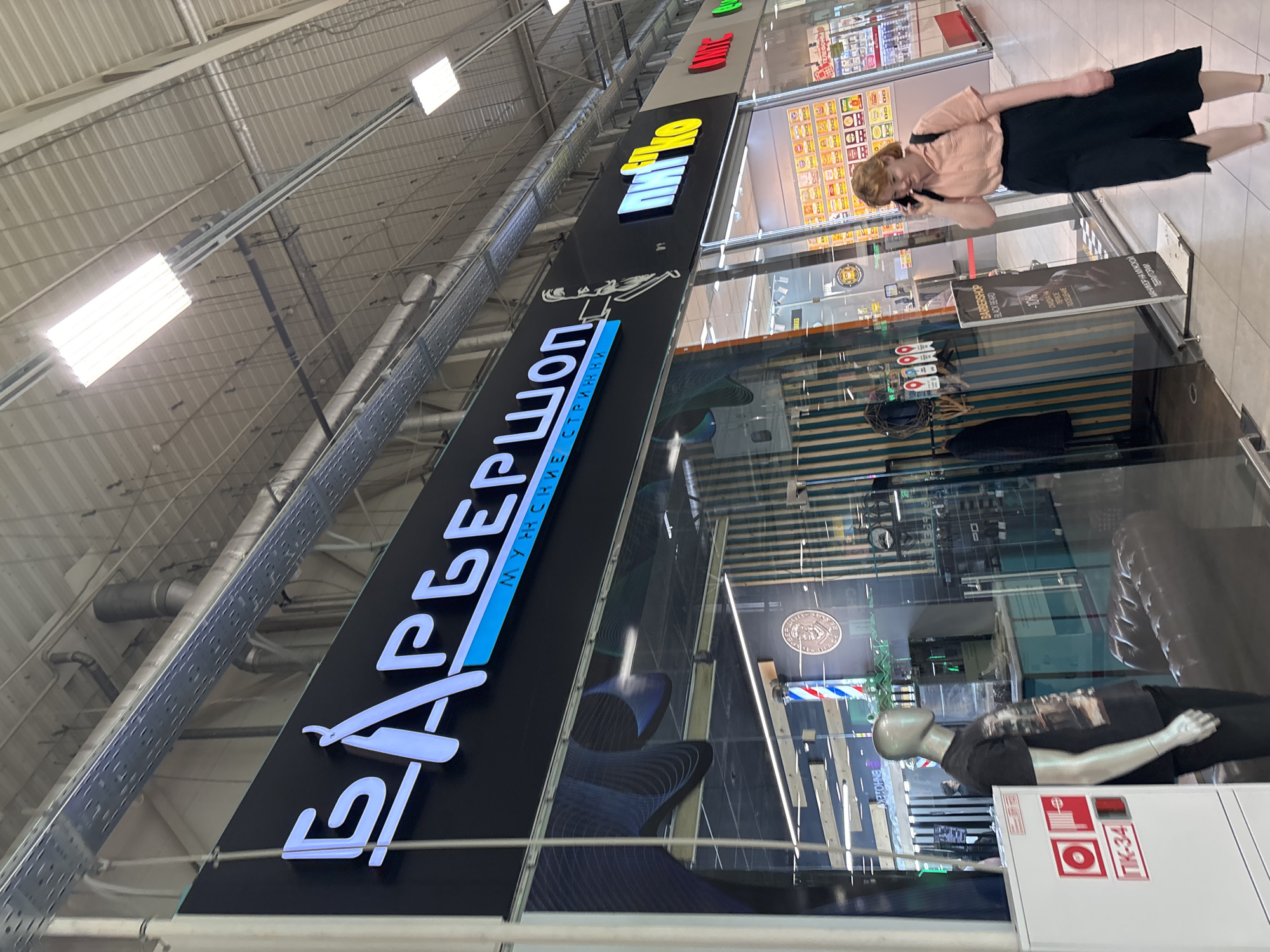 This full-length, vertically oriented photograph depicts an indoor scene within a shopping mall or commercial building, focusing on the storefront of a barbershop. The perspective is slightly angled upwards, looking down a brightly lit corridor.
The primary subject is a barbershop, prominently featuring a large, black illuminated sign with white and blue Cyrillic text that reads "БАРБЕРШОП" (Barbershop) and "МУЖСКИЕ СТРИЖКИ" (Men's Haircuts) below it. A stylized razor forms part of the barbershop's logo on the sign. Above this, another sign is visible with yellow and white text "ФИНО" (FINO), next to an unidentifiable red sign. The storefront itself is made of glass, reflecting parts of the interior, including a classic barber pole, a circular emblem on the wall, and what appears to be a black leather sofa. A mannequin, dressed in a dark t-shirt and black pants, stands inside the entrance, seemingly displaying clothing.
In the foreground, an A-frame sign stands outside the barbershop. Visible text on this sign includes "BARBERSHOP", "BLACK BEARD", "-10%", "CUIDA HA", "ПОСЕЩЕНИЕ", and "МАНИКЮР НА МУЖСКОЙ ТЕРРИТОРИИ" (Manicure in men's territory). On a white box near the mannequin, "ПК-34" is visible, likely a fire safety designation.
A woman is captured in motion on the right side of the frame, walking past the storefront while engaged in a phone conversation. She has reddish-blonde hair and is wearing a light-colored top and dark culottes.
The ceiling of the mall features a corrugated metal structure with numerous fluorescent light fixtures providing bright, artificial illumination. Exposed ducts, pipes, and cable trays are also visible overhead. The floor is tiled in a neutral color. The overall atmosphere suggests a typical modern indoor commercial environment, likely during daytime hours. All visible text is in Russian, confirming the implied location in Russia.