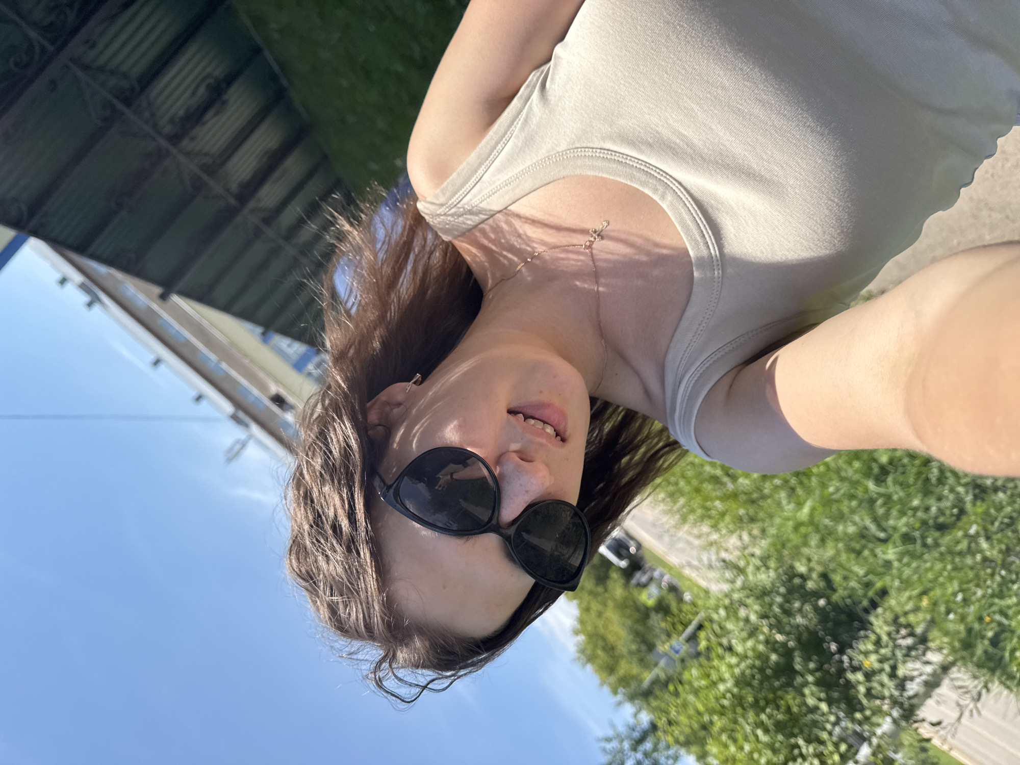 In Vereshchagino, Russia, a young woman with brown hair and black sunglasses is taking a selfie outdoors on a sunny day. She is wearing a light-colored tank top and a delicate necklace. Her hair is slightly windblown. Behind her, a paved area is visible, bordered by lush green foliage. In the background, a building with multiple floors and windows can be seen. The sky is clear and blue.