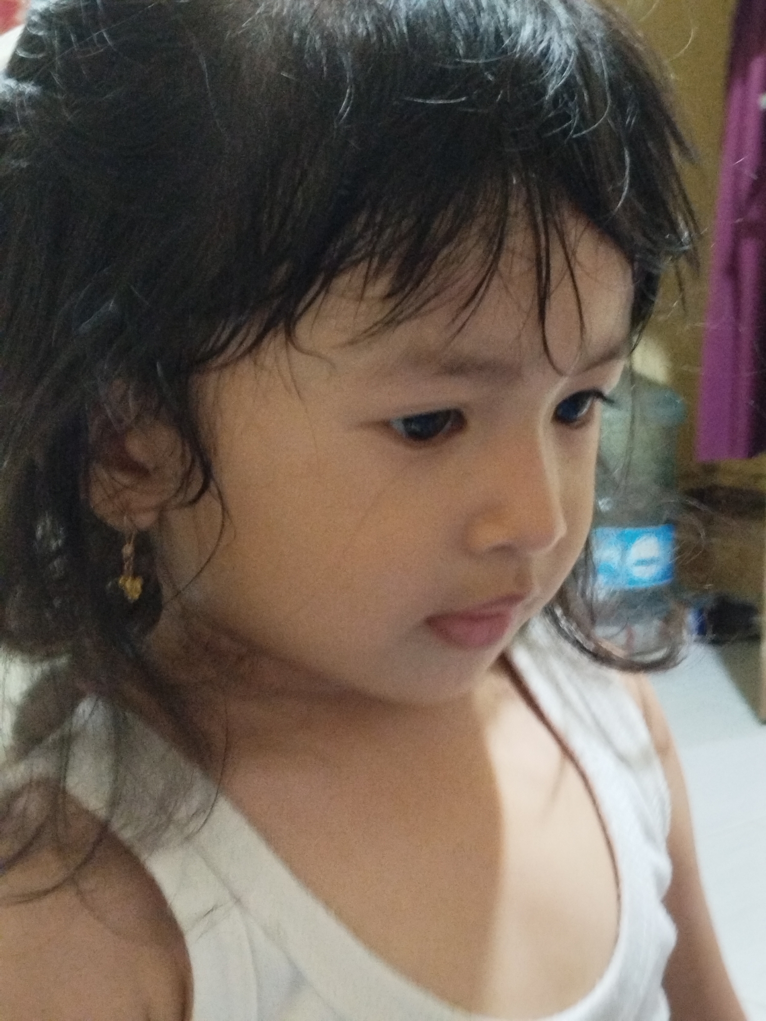 A close-up shot depicts a young child with dark, slightly disheveled hair, wearing a white tank top. The child has a serious expression and is looking down and to the right. A gold earring is visible on the child’s left ear. In the blurred background, a water bottle and a purple curtain can be partially seen. The location is unknown, and it is not possible to determine if the image was taken in Lhokseumawe, Indonesia from the given context.