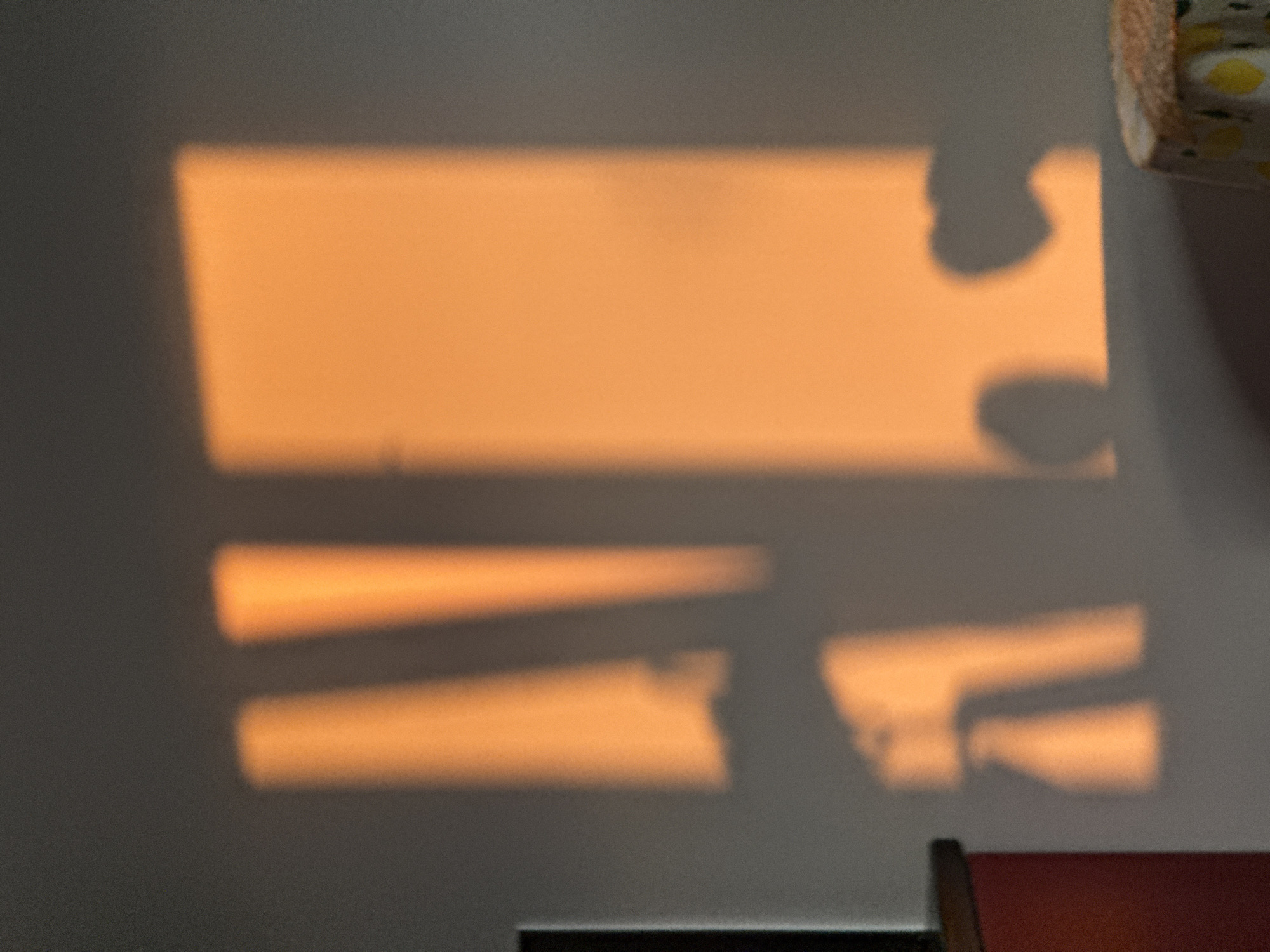 The image shows sunlight filtering through window blinds and casting shadows on a wall. There are two distinct sets of shadows: a large rectangular shape at the top with curved indentations, and several horizontal lines below it. A corner of a wooden object is visible in the lower right corner. A cylindrical object with a patterned fabric around it is present in the upper right corner. The wall surface appears to be a matte gray color. The location, Vozdovac Urban Municipality, Serbia, is not discernible from the image.