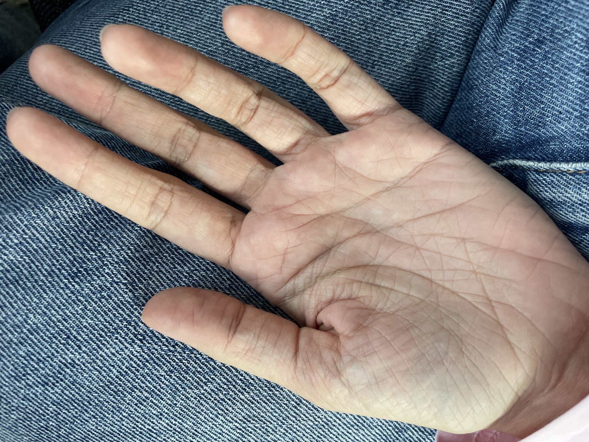 A close-up photograph depicts an open left human hand, palm upward, positioned against a textured blue denim fabric, likely clothing. The hand displays a light-to-medium skin tone, with visible creases and lines covering the palm and fingers. The fingers are slightly splayed. A portion of a light pink garment is visible along the lower left edge, near the subject's wrist. The hand appears to be at rest. The immediate environment consists of the aforementioned fabrics. No other subjects, objects, actions, or specific environmental features allowing for the identification of a city, such as Izhevsk, Russia, are present in the image.