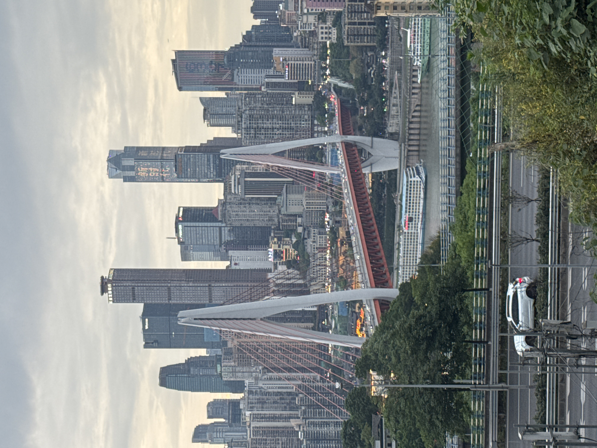 The image is a view of a cityscape in Nan'an District, China. The photograph features a modern bridge, the Chaotianmen Bridge, with white towers and a red roadway, spanning over a river. The skyline is composed of numerous high-rise buildings of varying heights and architectural styles. A portion of the riverfront can be seen with a ferry boat docked. In the foreground, there is an elevated road running parallel to the river. On the right side of the road, foliage is visible, along with a white automobile, possibly a police car. The sky appears cloudy. The overall tone is of an urban setting with modern infrastructure.