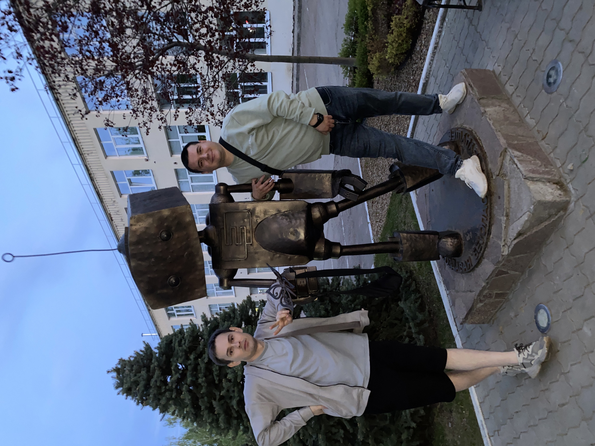 Two men pose with a metal statue in what appears to be an outdoor setting, possibly a park or public space. The man at the top is wearing a light green sweatshirt, blue jeans, and white sneakers. He has a black strap across his body. The man at the bottom is wearing a light gray sweatshirt, black shorts, and gray sneakers, and is giving the peace sign. They both appear to be interacting with the statue, which is set atop a stone base. The background features a building, trees, and paved ground. Based on the inscription on the stone base, the location is the city of Chaikovsky, Russia.