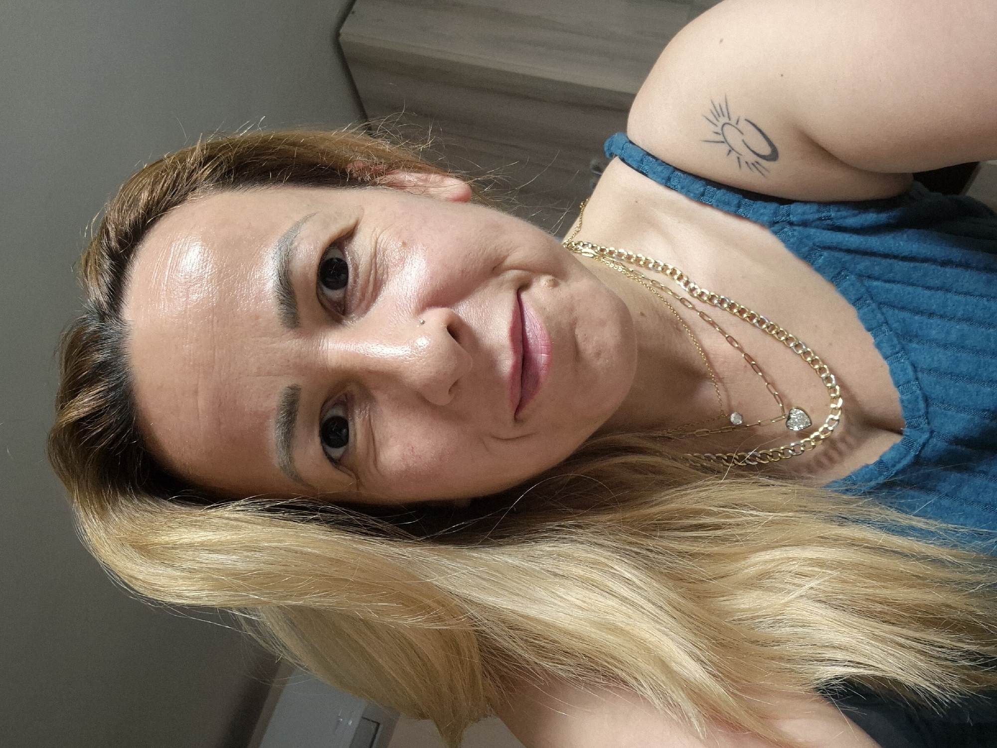 A medium shot shows a woman with long blonde hair and brown streaks, looking at the camera. She has fair skin, brown eyes, and is wearing a blue tank top, and gold necklaces. A tattoo of a sun and crescent moon is visible on her right shoulder. The background is out of focus, but appears to be an indoor setting with wooden cabinets.