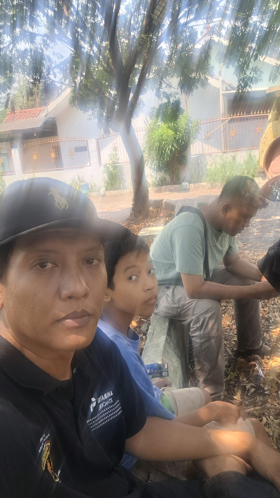 The image captures a candid moment of three individuals outdoors, likely in the city of Cilaku, Indonesia, based on the provided context. In the foreground, a man wearing a black baseball cap with a horse logo is taking a selfie. Beside him, a young boy with dark hair looks towards the camera with a curious expression. To the right, another man is seated, wearing a light green t-shirt and khaki pants, with his head bowed as if in thought or conversation. The background reveals a residential setting with a house featuring a tiled roof and a decorative metal fence. A tree with dense foliage casts dappled sunlight across the scene, illuminating the ground covered in dry leaves and twigs, suggesting a warm, possibly dry season. The overall atmosphere is informal and relaxed.