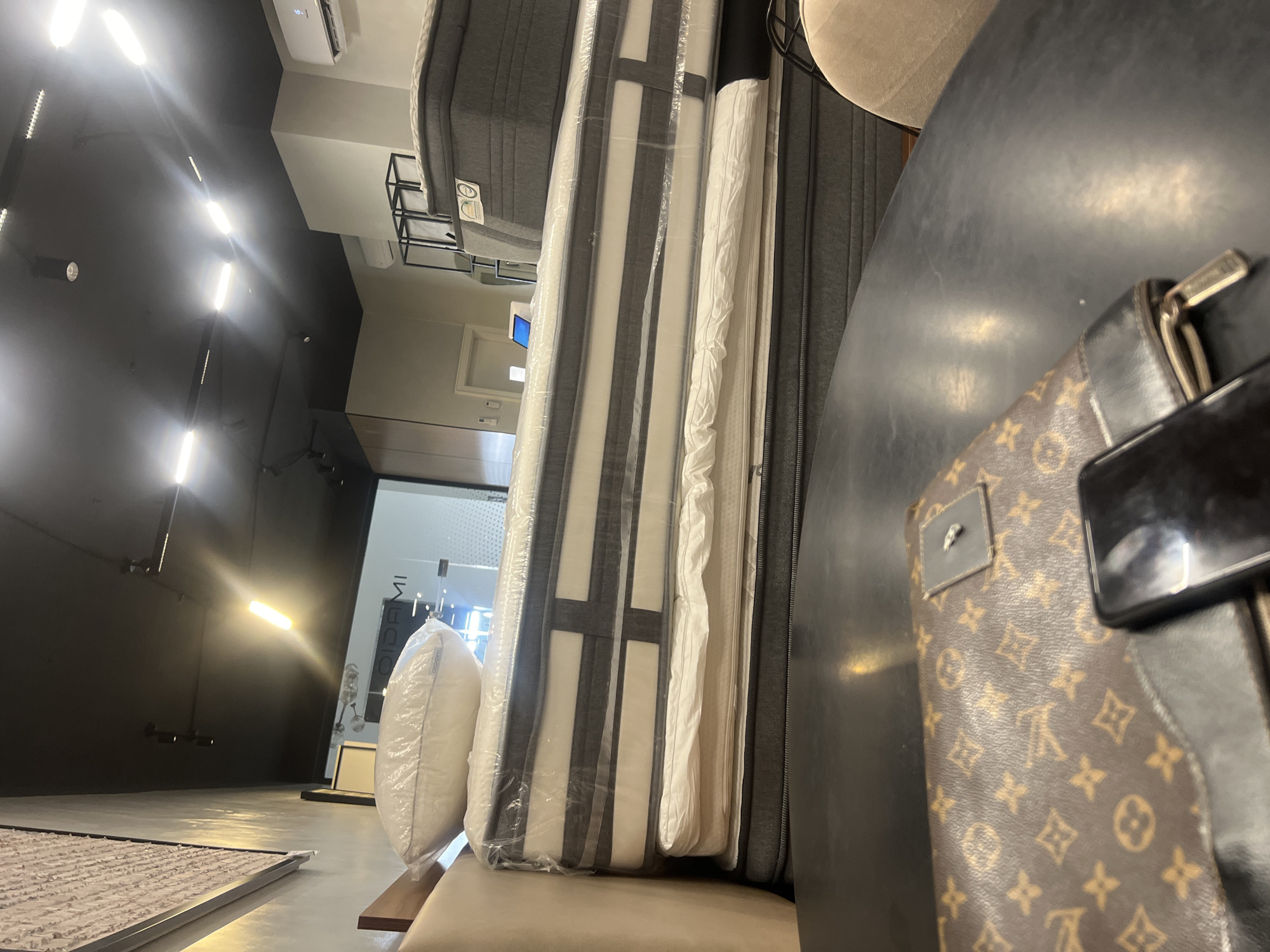 This is a photograph taken from a medium-low angle, looking across a dark, reflective surface, possibly a table, towards a stack of mattresses in what appears to be a retail store. In the foreground, a brown and gold patterned clutch or wallet with a silver clasp is visible, alongside a black smartphone. The mattresses are stacked high, with the top one wrapped in clear plastic and topped with a white pillow, also in plastic. The mattresses themselves have dark gray and white striped patterns.
In the background, the store interior is dimly lit with exposed ceiling lights and track lighting. The walls appear to be a neutral color, and there is signage visible that reads "OIDAMI." Further back, shelving units and a door can be seen. The overall impression is of a modern retail environment, likely a mattress or furniture store. The lighting suggests it might be evening or an indoor space without natural light. The presence of air conditioning units mounted on the wall indicates a climate-controlled environment. The photograph does not depict any specific identifiable landmarks or features that would place it definitively in Abuja, Nigeria, nor does it show any people or any interactions. The scene is static and focused on the display of goods.