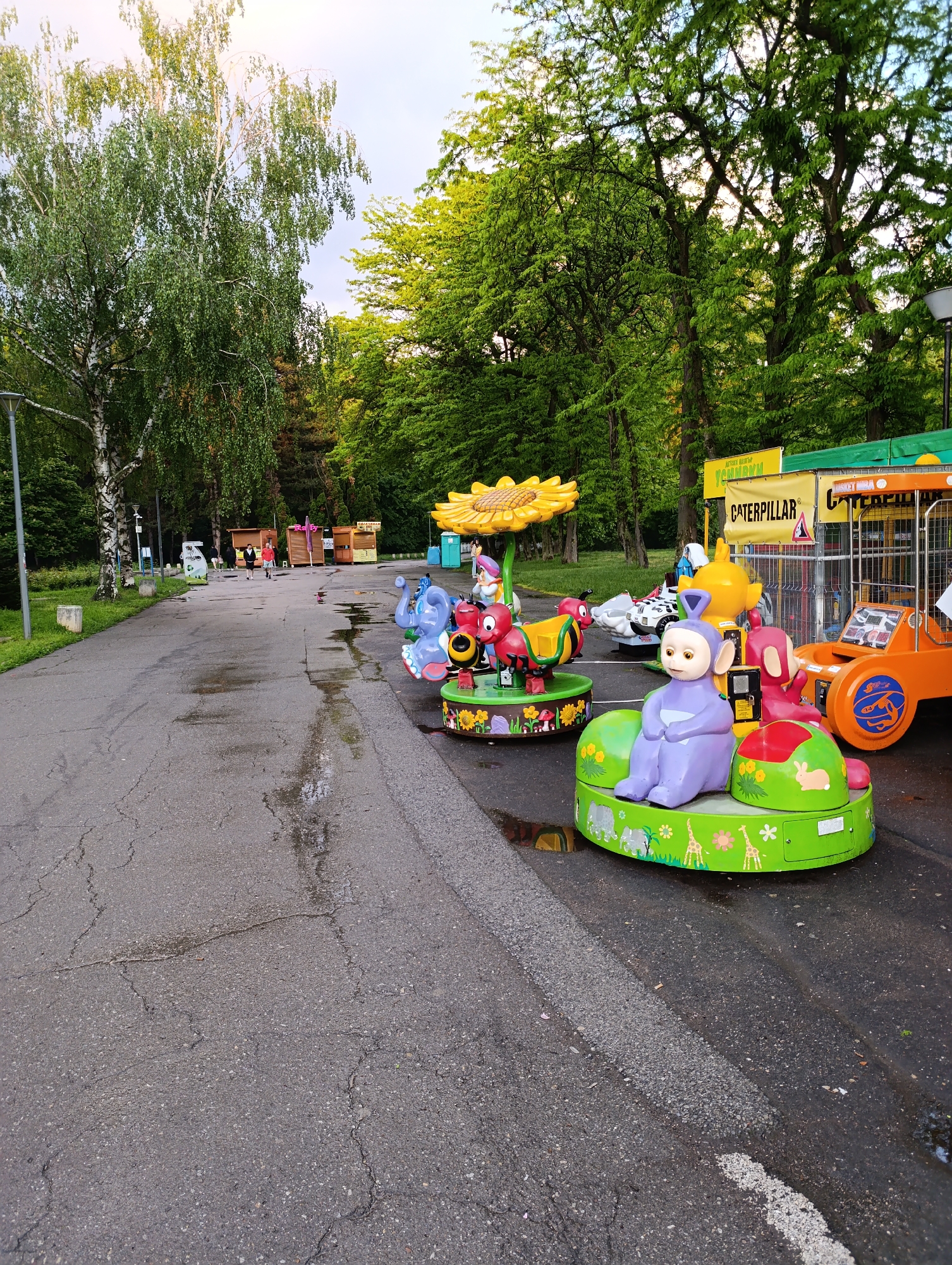 The image depicts a street scene in what appears to be a park or recreational area. The location is Sofia, Bulgaria, as indicated by the Cyrillic text on the "Caterpillar" sign. A variety of colorful kiddie rides, including a Teletubby figure, an elephant, a sunflower, an ant, and a small orange vehicle, are lined up on the edge of the asphalt road. The road itself is wet, suggesting recent rain. In the background, there are people walking along the road, and some small wooden kiosks or stalls are visible. Trees line the street, providing shade. The overall scene suggests a temporary or seasonal amusement setup within a public park.