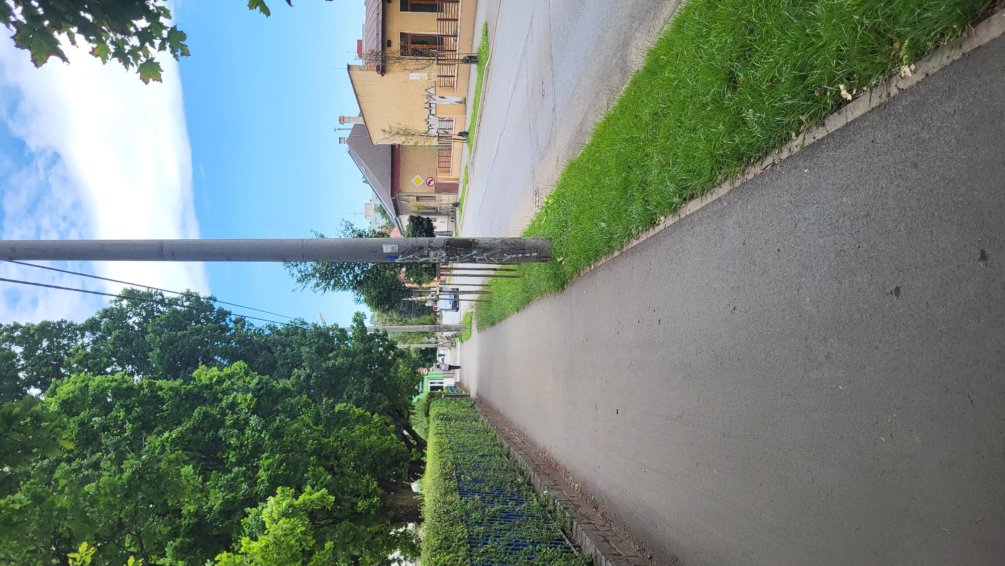 A paved pedestrian walkway runs along the left side of a two-lane asphalt road in Prešov, Slovakia. The walkway is bordered on the left by a blue-fenced hedge and mature trees. On the right, the walkway is separated from the road by a strip of green grass and a concrete curb. The road itself is lined with buildings on the right side of the frame, appearing to be residential or commercial structures. A single utility pole stands on the right side of the walkway, just before the grass strip begins. The sky is blue with scattered clouds. A single person is walking away from the camera on the walkway in the distance, and a dark-colored car is parked on the right side of the road, further down. The scene appears to be a quiet, suburban street.