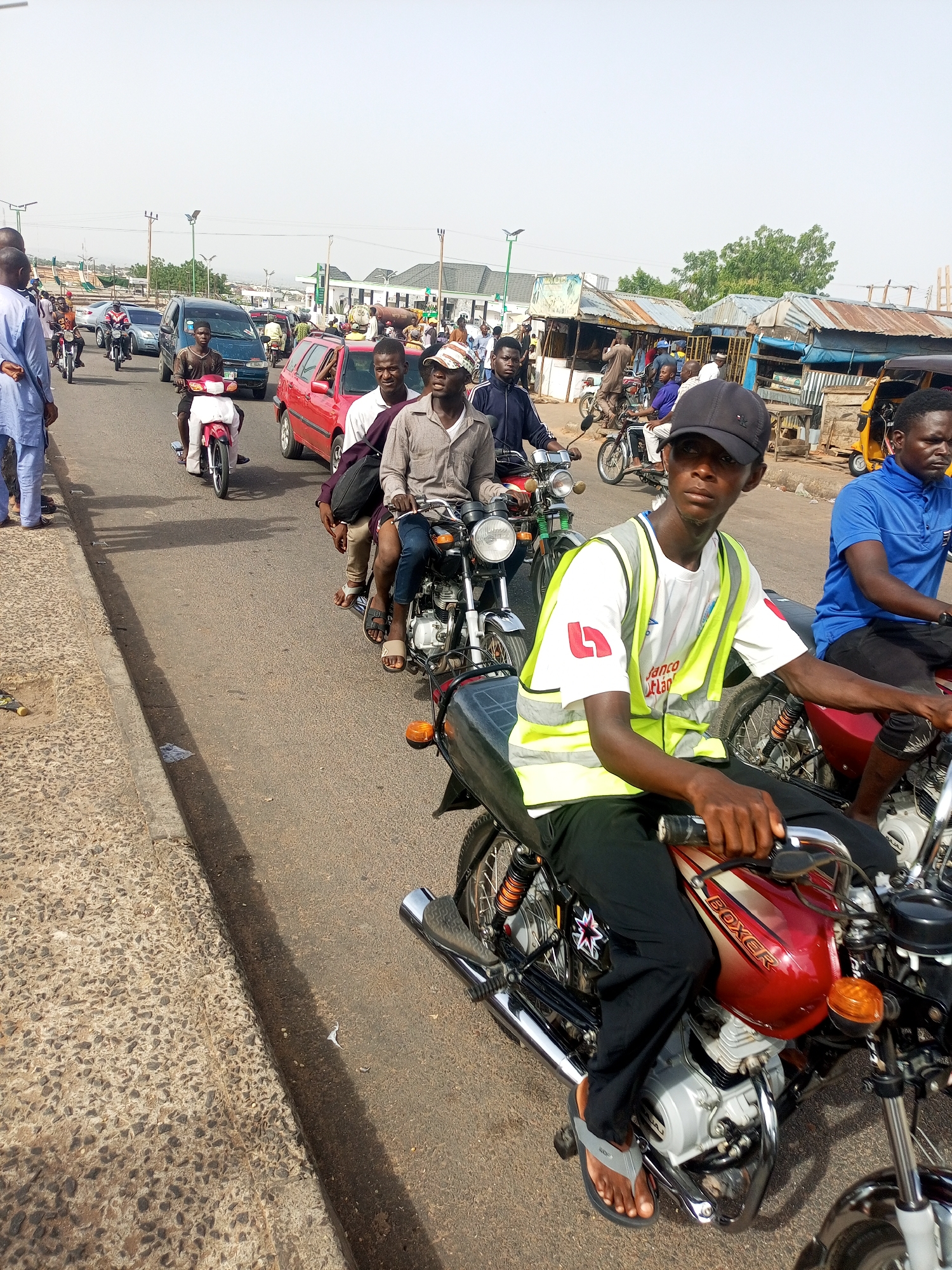 The image depicts a busy street scene in Gombe, Nigeria. A line of motorcycles, mostly with riders and some with passengers, is waiting in traffic. The riders appear to be mostly young men, casually dressed. One rider in the foreground wears a reflective safety vest over a t-shirt and a baseball cap. Some riders are wearing sandals.
Behind the motorcycles are several cars and other vehicles. Pedestrians are visible on the sidewalks and near the street. There are single-story buildings and shops along the roadside, with corrugated metal roofs. The sky is bright, suggesting a sunny day. The infrastructure appears basic and somewhat crowded, characteristic of urban areas in Nigeria.