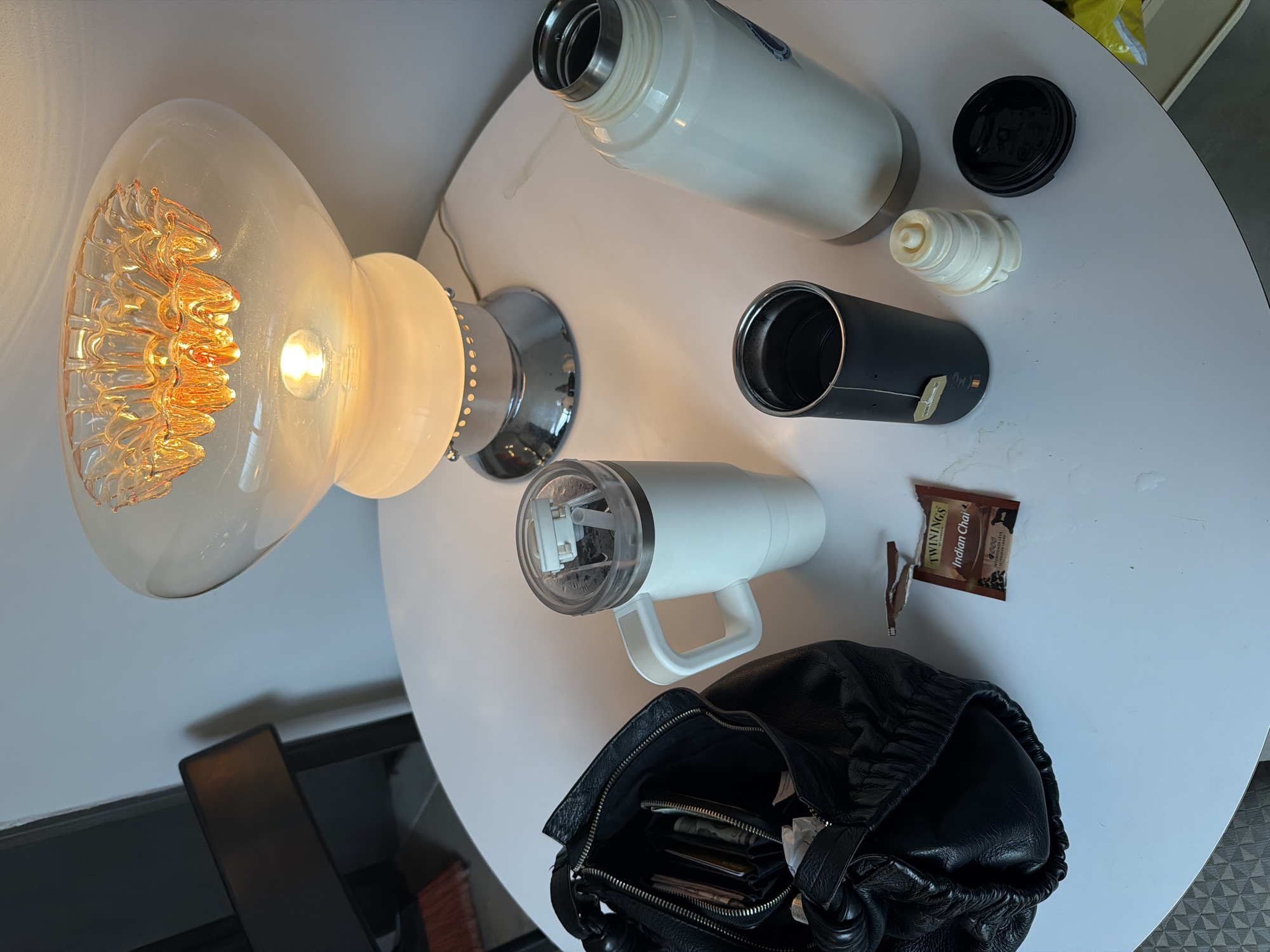 A high-angle shot depicts a collection of items on a white, round table. The objects are a lamp with a clear, decorative glass shade, a white thermos, a black tumbler with a tea bag, the parts of the thermos cap, a white mug with a handle, and a black leather handbag. A Twinings Indian Chai tea bag wrapper lies near the handbag. In the background, part of a black chair can be seen, along with a section of a white wall. The scene does not provide any discernible clues about a specific location in Buenos Aires, Argentina.