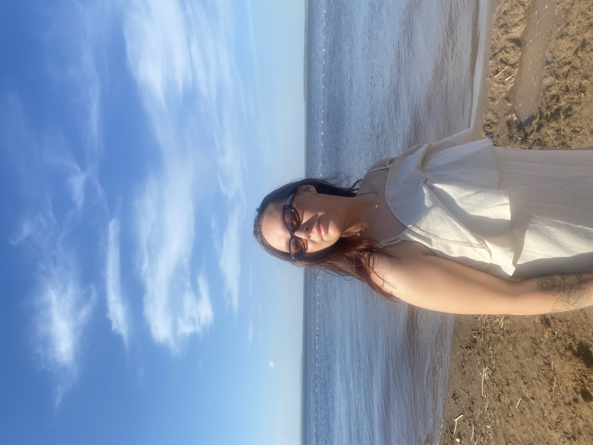 A woman stands on a beach, waist-deep in water. She wears a white dress and sunglasses. The sky is blue with scattered clouds. The location is Chaikovsky, Russia.