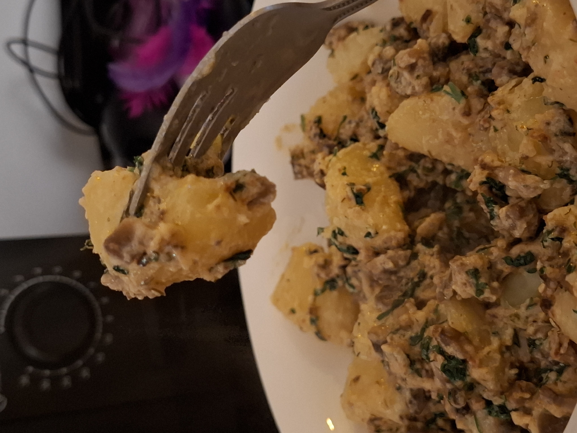 A fork is lifting a portion of diced potatoes mixed with ground meat and herbs from a white bowl. The dish appears to be creamy and seasoned. In the background, out of focus, is a dark appliance with a circular dial and an object with purple and pink elements. The overall scene suggests a casual mealtime setting. There is no identifiable information about the location in Zemun Urban Municipality, Serbia.