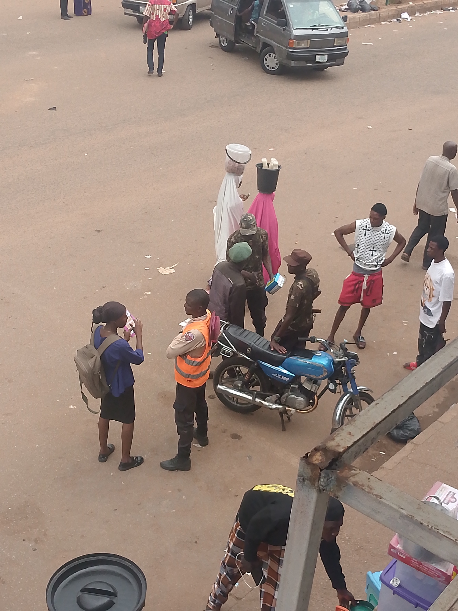 The image depicts a street scene in Bukuru, Nigeria. Several people are visible. A young woman with a backpack is taking a picture with her phone. A young man wearing an orange vest stands next to her. Two uniformed men, possibly military personnel, are near a blue motorcycle. One is sitting on the motorcycle. Two individuals carry goods on their heads, one with a white bucket and head covering, the other with a black bucket. Two other young men stand nearby. A gray van is parked in the background. A person in a red shirt and jeans walks away from the camera. Another man walks in the background. Part of a rusted metal structure is visible in the lower right corner of the image. A black trash can is visible in the lower left.