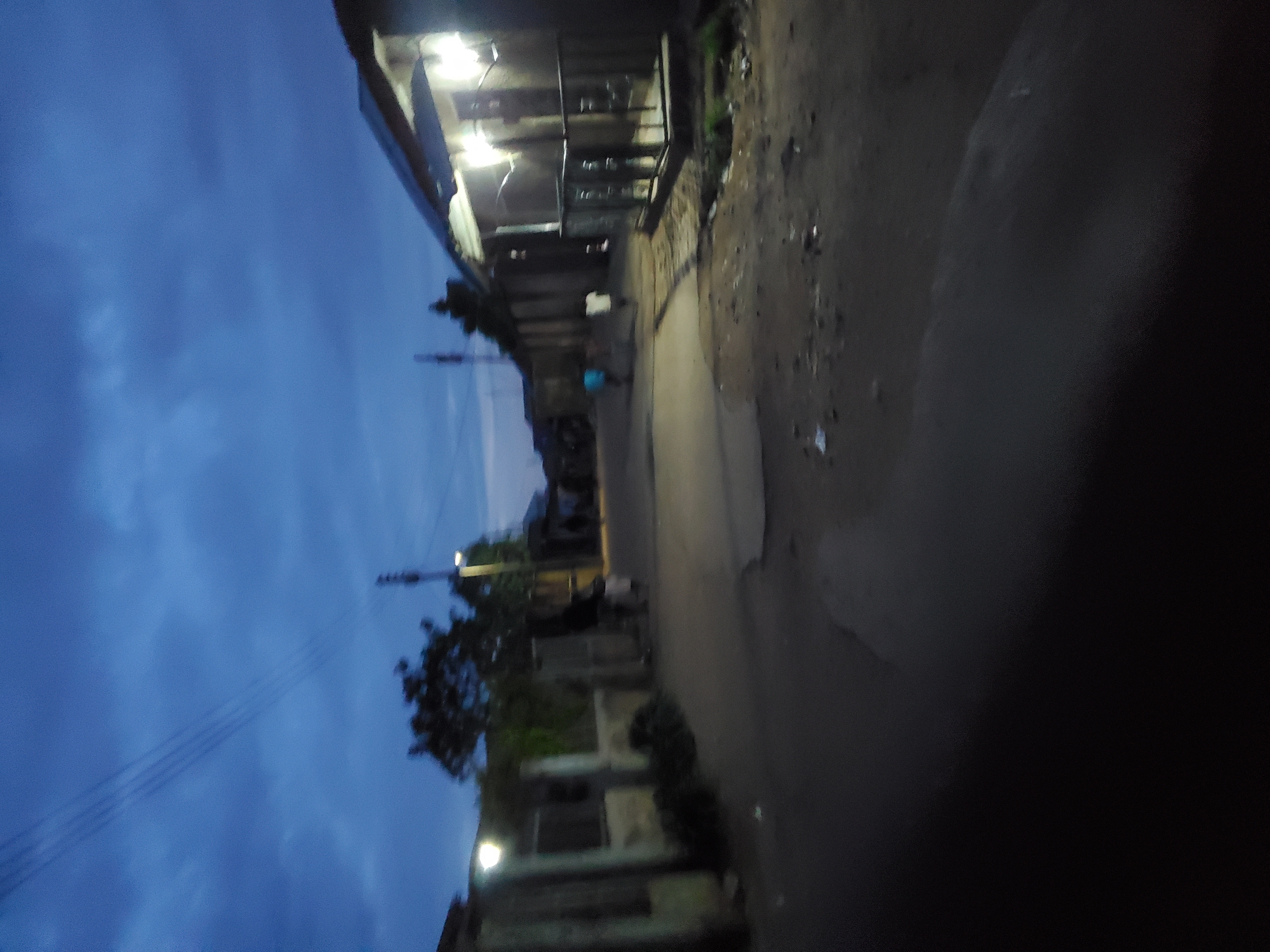 The image depicts a street in Jos, Nigeria at dusk. The street is lined with buildings, some with lit windows. The road surface is uneven and appears to be concrete or asphalt. A figure can be seen further down the street, although details are obscured by the lighting conditions. Utility poles and overhead wires are visible against the evening sky, which is a gradient of blue. Vegetation, including a tree, is located along the street. The overall atmosphere is dim.