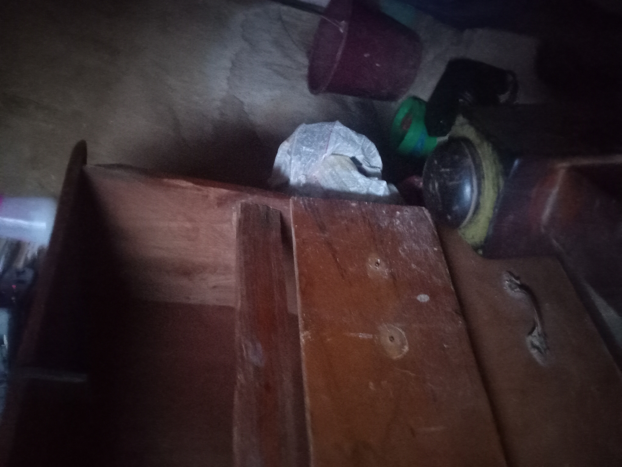 The image depicts an interior scene, likely a storage area or a room in a dwelling, located in Bukuru, Nigeria. A wooden cabinet with multiple drawers is prominent, showing signs of age and wear. One drawer, in particular, has a handle that is either broken or in a state of disrepair. Beside the cabinet, a dark-colored bucket, possibly made of plastic, is visible. To the left of the bucket, two smaller, brightly colored containers, one green and one red, are present, suggesting they might be used for storage or containing items. A crumpled white material, possibly plastic sheeting or a bag, is situated behind these containers. In the foreground, a dark, rounded object with a metallic top rests on a textured, yellow surface, which could be a lid for a pot or a similar item. The background wall appears to be made of rough, textured material, possibly concrete or plastered stone, consistent with common building materials in the region. The lighting in the scene is dim, casting shadows and obscuring some details, creating an overall impression of a utilitarian space. There are no people visible in the image.