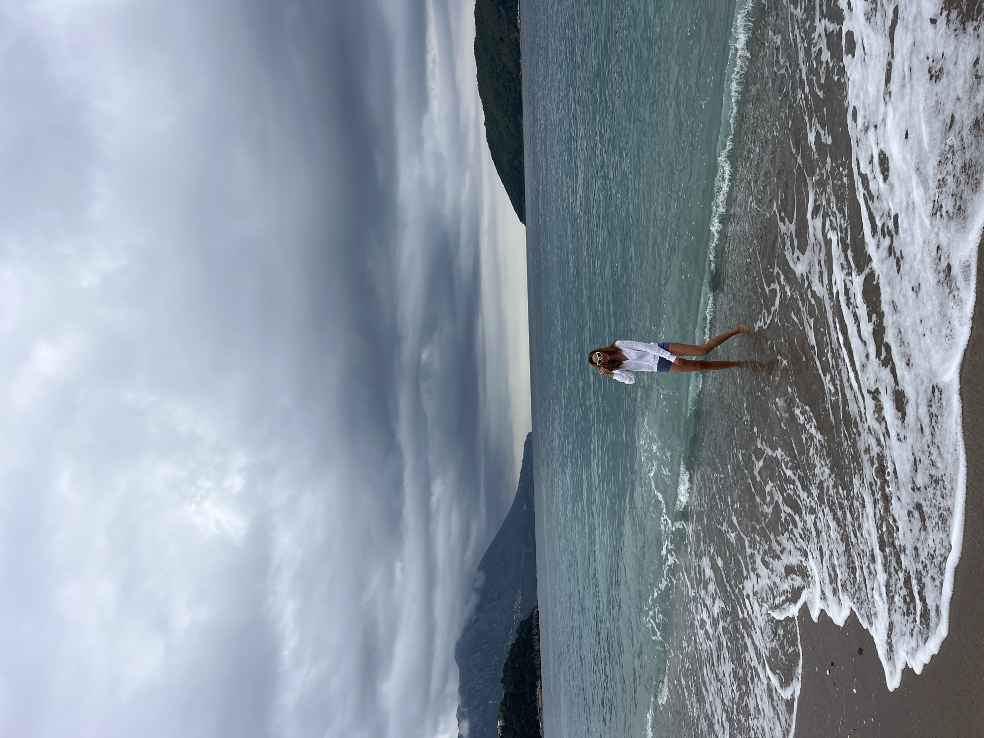 The image, oriented horizontally, depicts a serene coastal landscape on an overcast day. In the foreground, a woman stands in the shallow, clear blue-green sea water, facing slightly towards the viewer. She has long brown hair and is wearing sunglasses, a white long-sleeved shirt, and blue denim shorts. Her bare feet are visible, submerged where gentle white foamy waves meet the dark sandy shore.
The calm sea stretches across the midground, displaying a subtle texture on its surface. In the background, majestic dark green and grey mountains rise dramatically from the coastline, their peaks partially veiled by thick, low-hanging grey clouds. The sky above is entirely covered with these dense, overcast clouds, casting a soft, diffused light over the entire scene. The atmosphere is tranquil and peaceful, with the rugged mountains providing a dramatic backdrop to the calm waters. There is no visible text in the image.