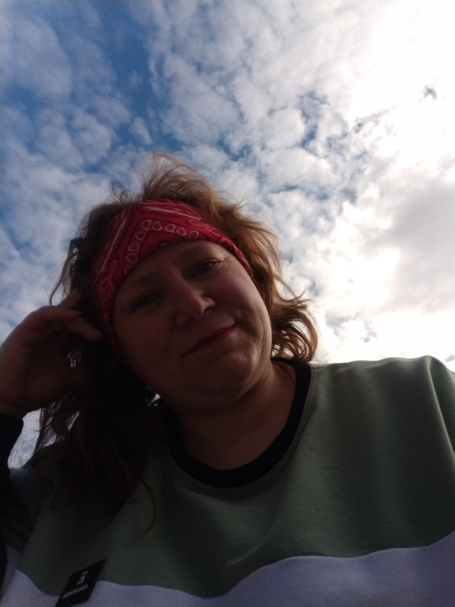 A low-angle selfie shows a woman with fair skin and auburn hair, wearing a red patterned bandana, a light green top with a black neckline, and a white lower section. She is looking down at the camera with a slight smile. Her left hand is raised near her head. The background consists of a blue sky with scattered white clouds. No specific location in Chaikovsky, Russia, can be determined from the image.