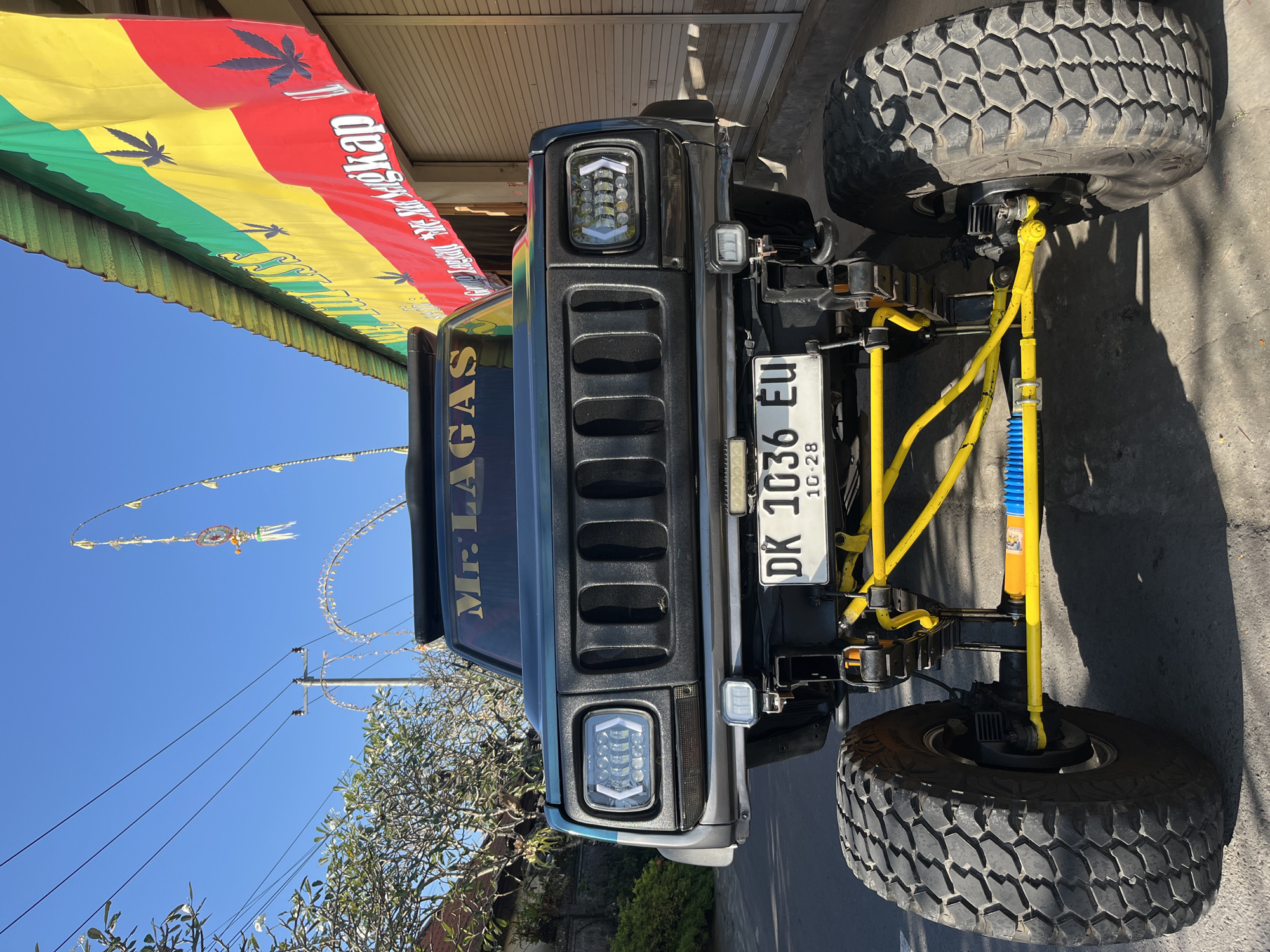 In Sukawati, Indonesia, a heavily modified, dark green off-road vehicle is parked on a paved street facing the camera. The vehicle features oversized, knobby tires, a lifted suspension with prominent yellow components, and custom headlights. The front grille is black and has a distinctive vertical slot pattern. A license plate reading "DK 1036 EU" is visible below the grille. Above the windshield, the words "Mr. LAGAS" are displayed in gold lettering. To the right of the vehicle, a building with a metal roof is visible, adorned with a large banner in red, yellow, and green stripes, featuring cannabis leaf imagery and text in Indonesian. The sky is clear and blue, with some electrical wires and trees on the left side of the frame. Traditional Balinese decorative flags are also present.