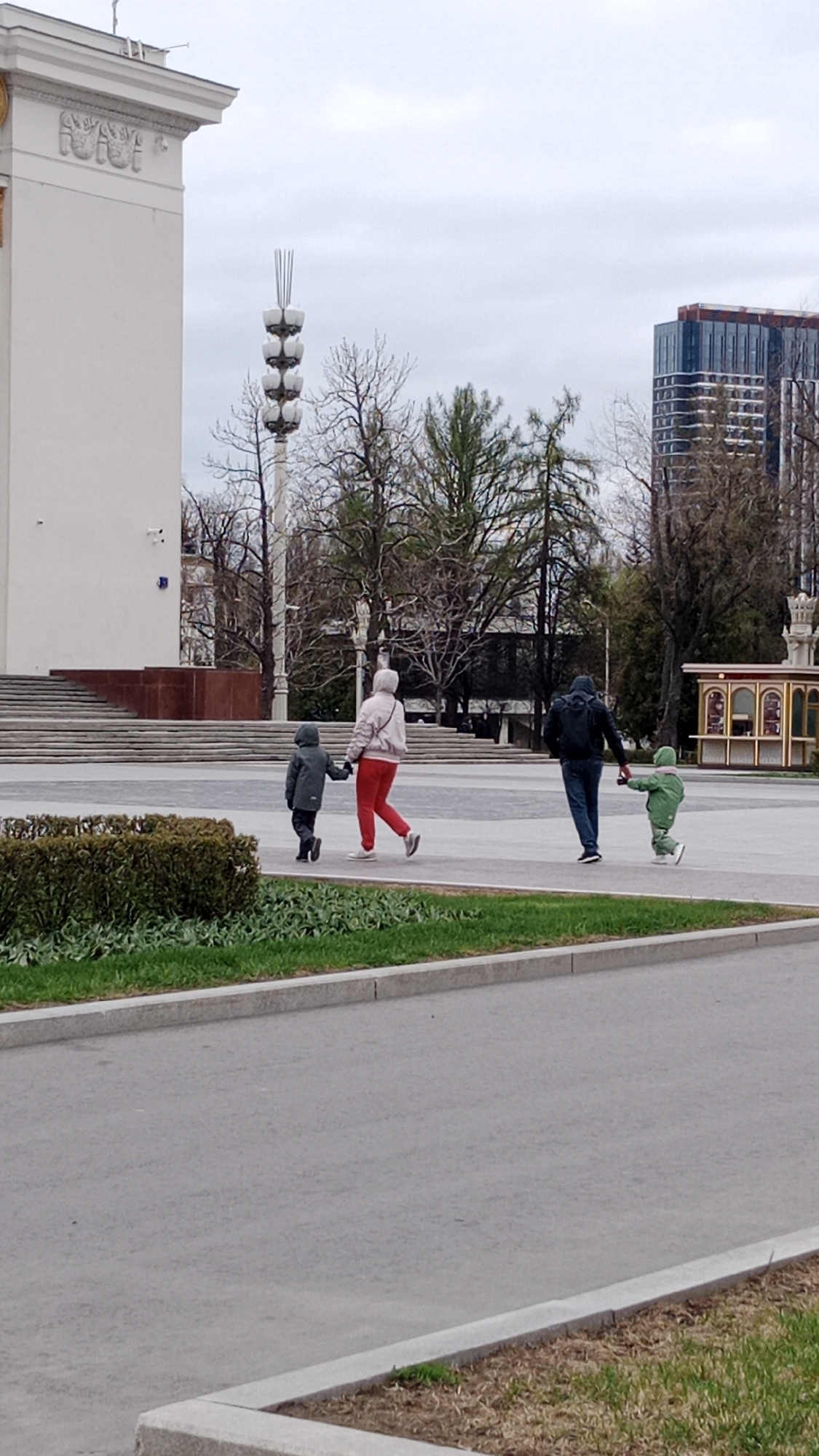 A family of four walks across a paved plaza on an overcast day in Moscow, Russia. To the left stands a large, white building with decorative architectural elements, adjacent to a series of stairs. Further back, a light post with multiple bulbous lamps extends above bare trees. A modern high-rise building is partially visible behind the trees. The family consists of a woman in a light pink jacket and red pants holding the hand of a child in a dark gray coat. A man in a dark jacket holds the hand of a child in a bright green snowsuit, who appears to be running or being pulled along. A low hedge lines the edge of the plaza.