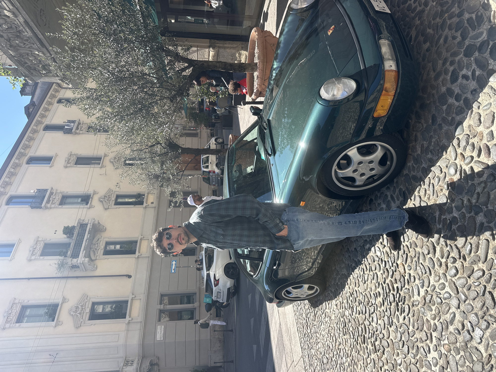 The image depicts a man standing next to a green Porsche car on a cobblestone street in Milan, Italy. The man is wearing a plaid shirt, jeans, and sunglasses. The car is parked near a building with ornate windows and balconies. People are visible in the background. The scene is bathed in sunlight. A street sign for "VIA MARCONI" is visible.