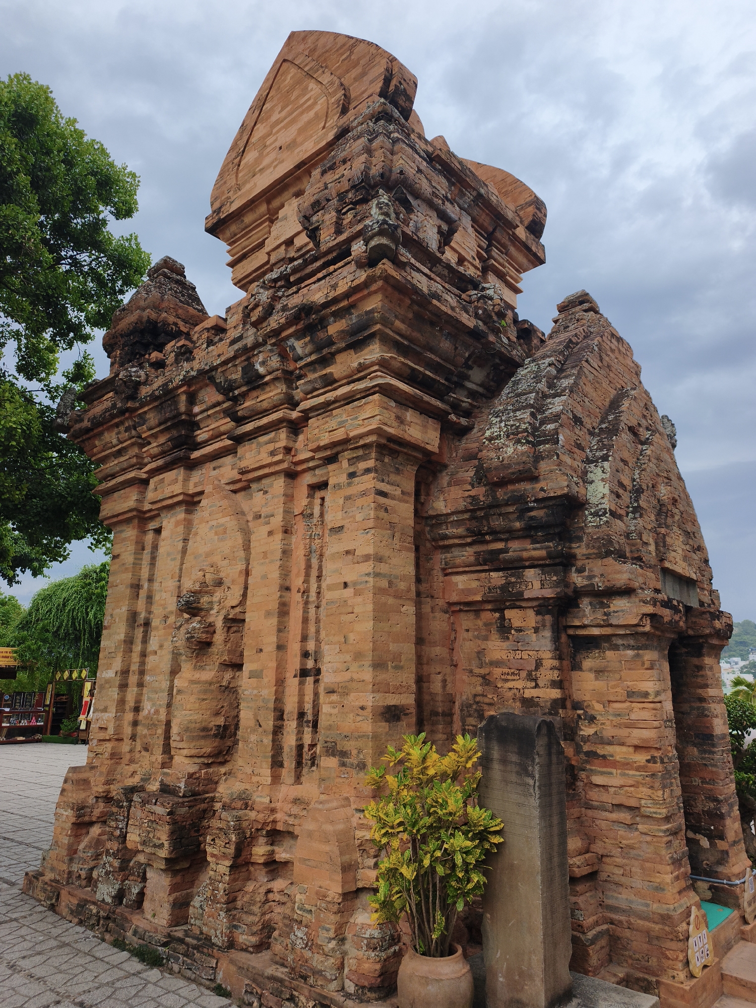 The image depicts the Ponagar Cham Towers, a historical complex located on a hill in the city of Nha Trang, Vietnam. The primary subject is one of the brick towers, constructed in the traditional Cham architectural style. The tower is made of reddish-brown bricks, showing signs of age and weathering, with some areas displaying green moss and dark discoloration. The structure has multiple tiers and elaborate decorative elements carved into the brickwork, including what appear to be figures or patterns.
In the foreground, a potted plant with vibrant yellow and green leaves stands to the left of a tall, upright stone stele. The ground in front of the tower is paved with grey stone tiles. To the left of the tower, a lush green tree provides shade. In the background, the sky is overcast with grey clouds, and a glimpse of distant buildings and greenery can be seen. There are no people visible in the image.