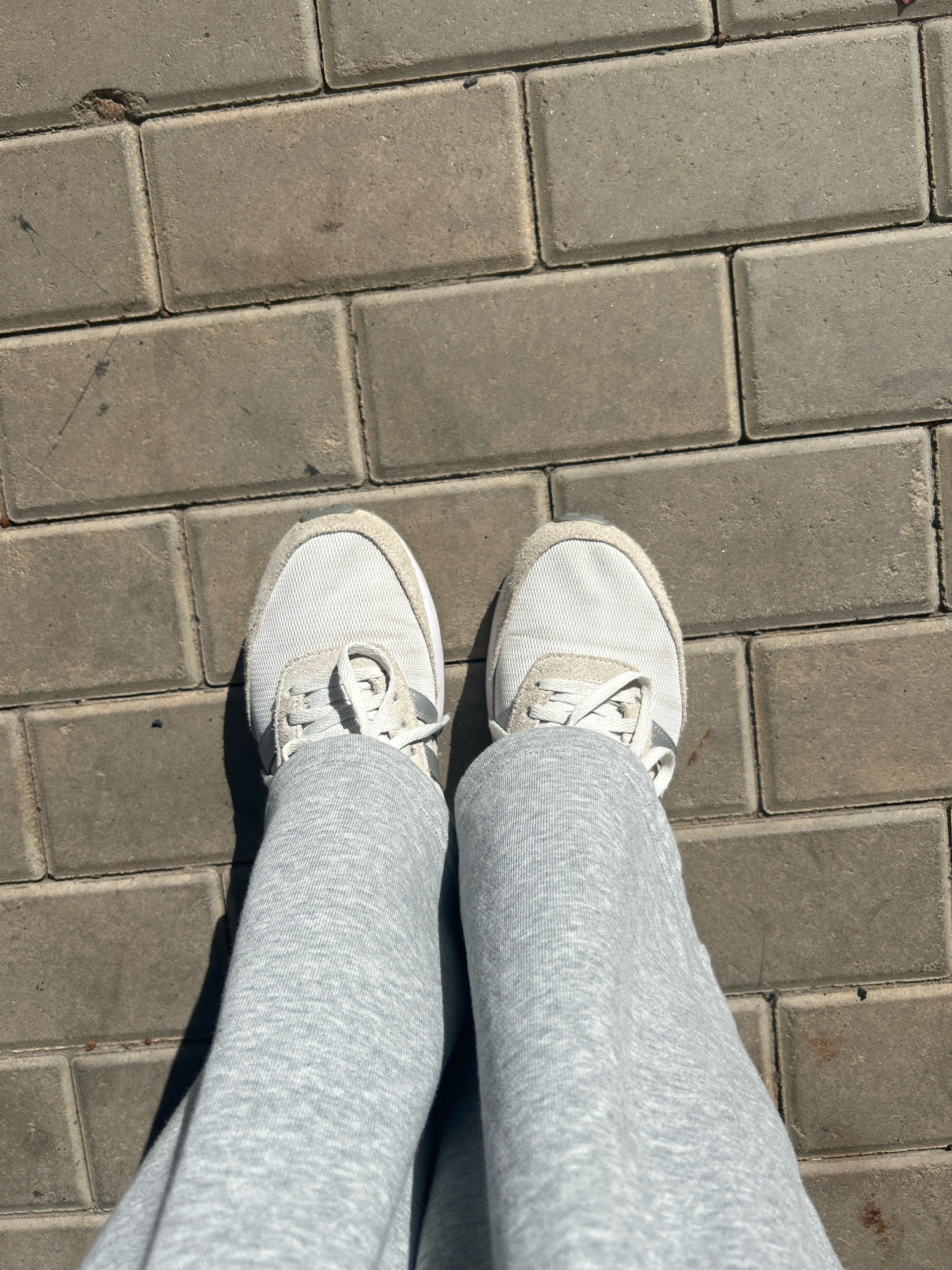 A pair of white athletic shoes with grey laces are worn by an individual standing on a sidewalk paved with rectangular grey bricks. The individual is wearing light grey sweatpants. The scene is outdoors, under natural sunlight, casting a shadow from the person's legs to their left. The cobblestone sidewalk appears to be slightly worn with some minor debris visible. The image is taken from a top-down perspective.